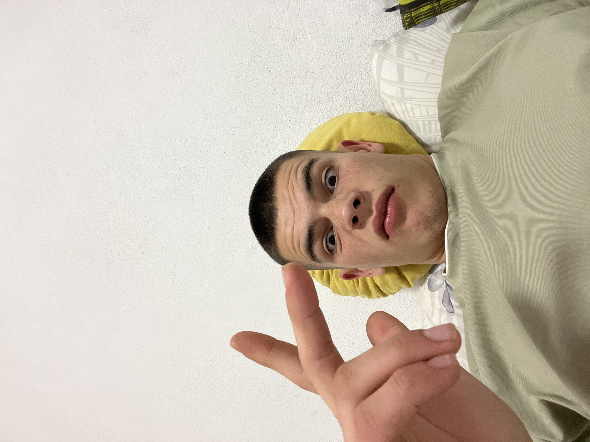 A young adult male is lying on his back. He has short, dark hair and is making a hand gesture. The background includes a white wall and bedding with a yellow cushion. No information allows the location to be identified as Karavelovo, Bulgaria.