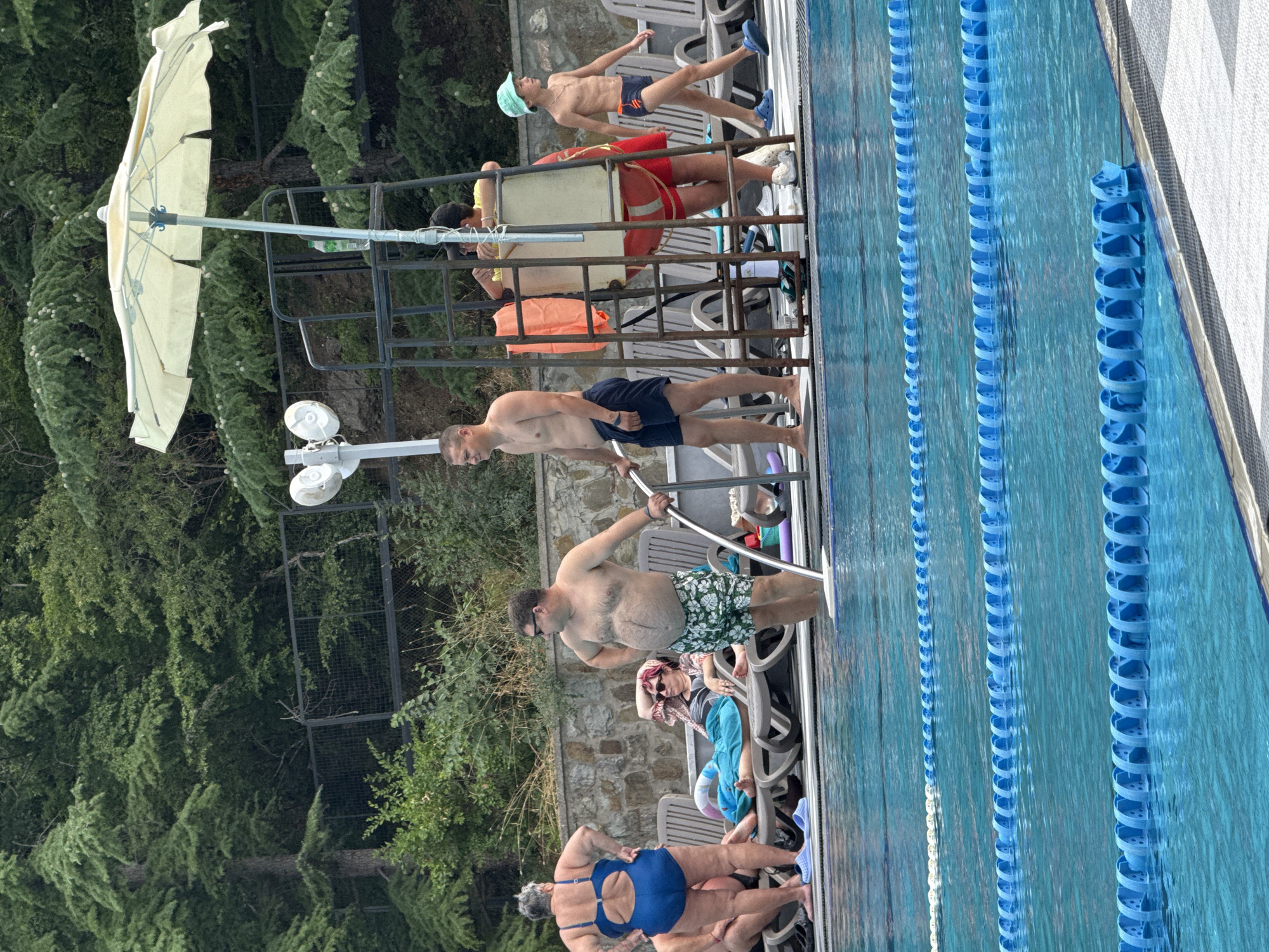 The image depicts an outdoor swimming pool area with several people. In the foreground, the bright blue water of the pool is visible with lane markers. Behind the pool, there is a stone wall and lush green trees.
Several individuals are present around the pool. On the left, a woman in a blue swimsuit stands near the edge of the pool. Next to her, a man in floral swim shorts stands partially submerged in the water. Further back, a woman in a patterned top and a turquoise towel is reclining on a lounge chair.
In the center of the image, a lifeguard chair is set up, equipped with a white umbrella. Several people are near the chair, including a man in dark blue swim trunks and a young boy in a light blue hat and swim trunks. An orange floatation device is visible attached to the lifeguard chair.
The scene appears to be a recreational area, possibly a resort or public pool, with a backdrop of dense foliage and trees, consistent with a location in or near Массандровский поселковый совет, Ukraine, given the vegetation and general setting.