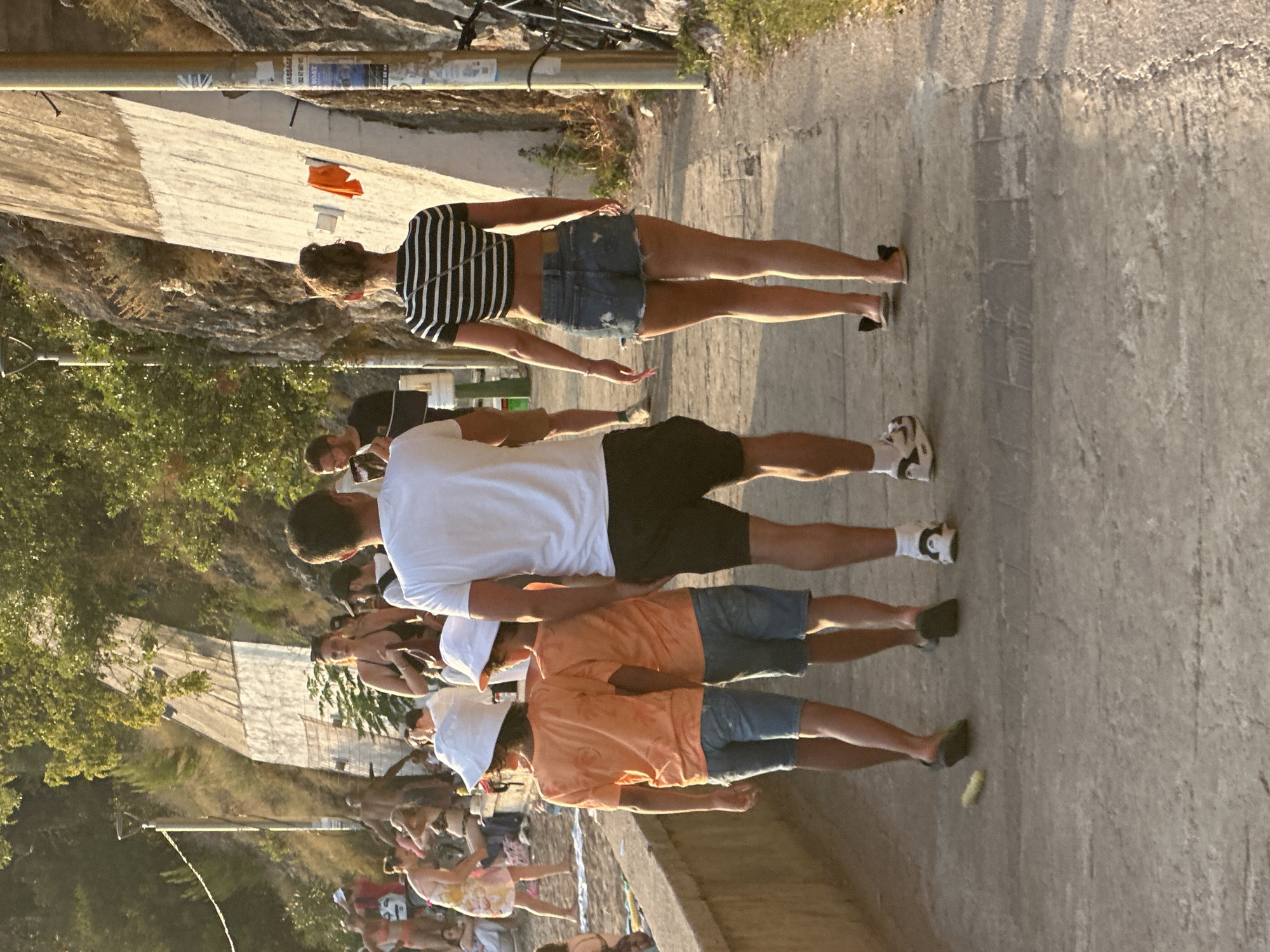 The image captures a scene on a paved walkway near the coast in Budva, Montenegro. In the foreground, two young boys wearing white bucket hats and orange t-shirts walk side-by-side, their backs to the camera, each with denim shorts. A man in a white t-shirt and black shorts stands to their right. Further ahead, a woman with her hair in a bun, wearing a black and white striped crop top and denim shorts, walks away from the viewer. The walkway is bordered by a low concrete wall on the left, and a steep, rocky embankment covered in vegetation on the right, with a concrete retaining wall. In the background, more people are visible on a sandy area that appears to be a beach. The lighting suggests it is late afternoon or early evening, with long shadows cast by the setting sun.