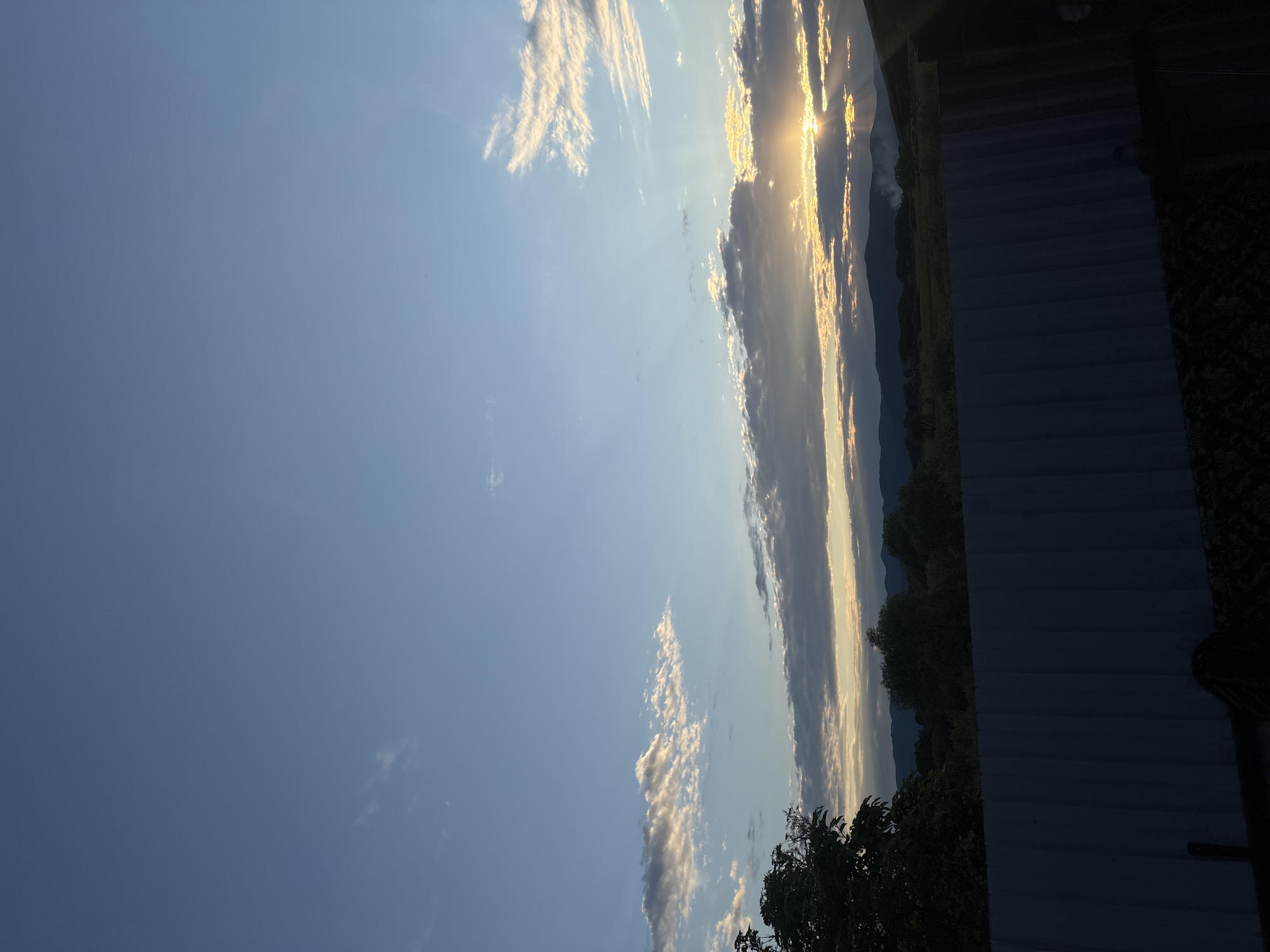 The image depicts a serene sunset over a rural landscape. The sky is a gradient of blue, transitioning to warm hues of orange and yellow near the horizon where the sun is setting. Wispy clouds are scattered across the sky, catching the golden light. In the foreground, a dark, corrugated metal fence runs horizontally across the bottom of the frame. Beyond the fence, rolling hills covered in lush green vegetation and trees are visible. The sun's rays pierce through the clouds, casting a dramatic light on the scene. A faint plume of smoke or mist rises from the distant landscape, adding an atmospheric touch. The overall impression is one of peace and natural beauty.