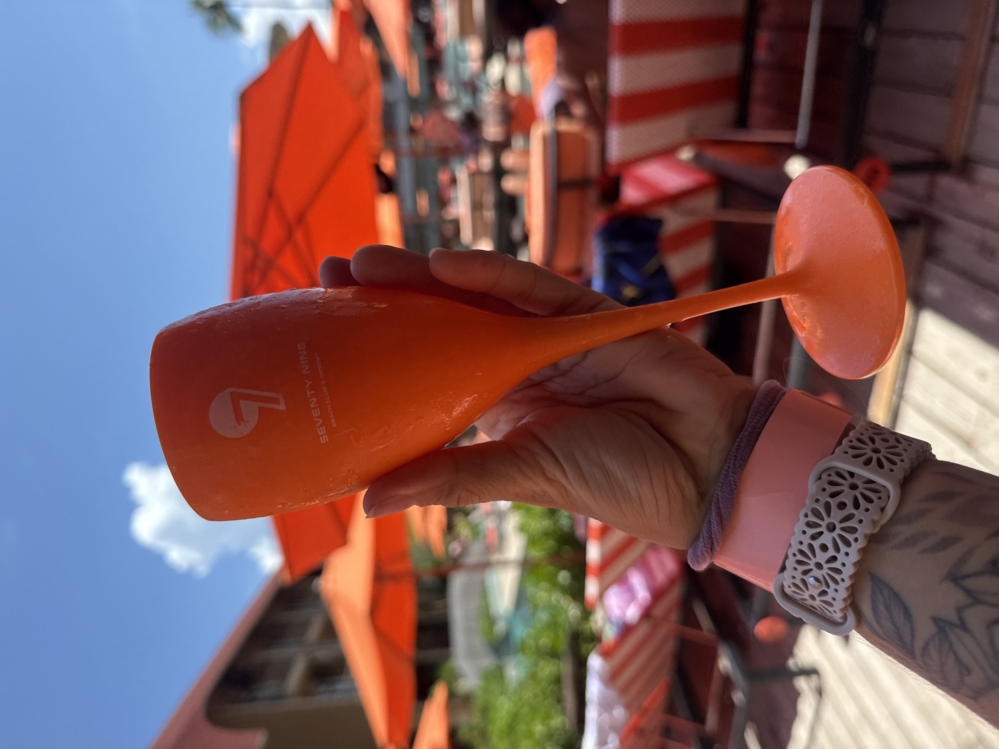 A close-up, eye-level shot shows a person's hand holding a bright orange champagne flute. The flute is frosted and has a white logo with the number 7 and the words "SEVENTY NINE BEACH CLUB & RESORT" printed on it. The hand has visible tattoos and is wearing two wristbands: a pink one and a lighter pink patterned one. The background is blurred, but shows an outdoor resort setting with orange umbrellas, lounge chairs with red and white stripes, and a swimming pool. The sky is blue with some clouds. The scene suggests a vacation or leisure activity in a tropical resort. The location is identified as Ko Samui District, Thailand.