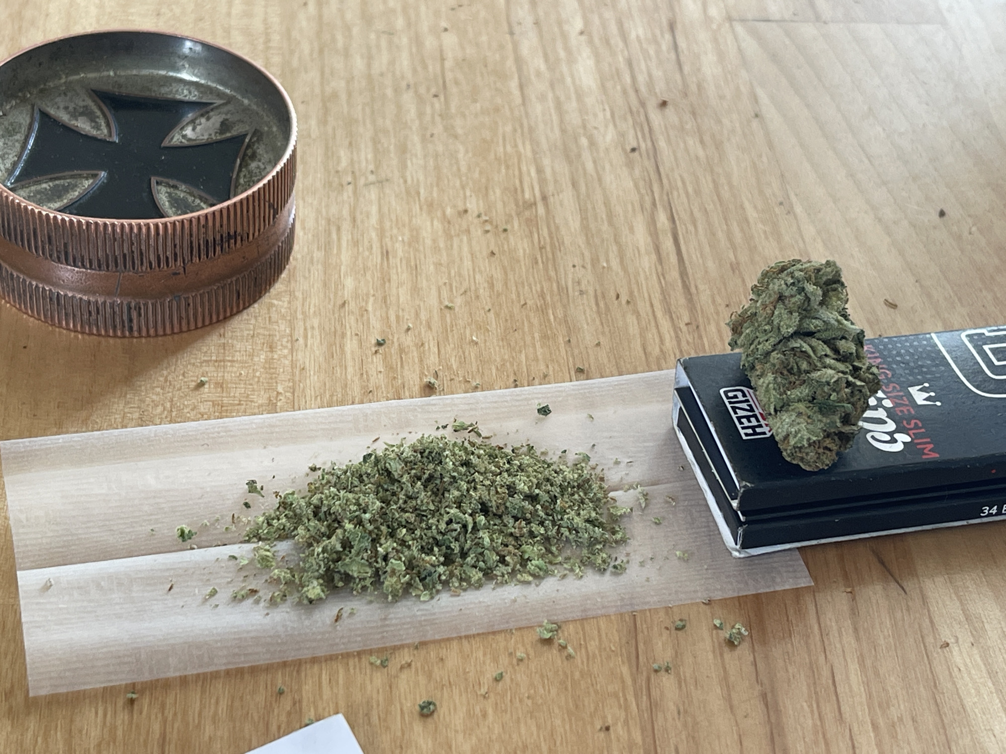 A copper-colored metal grinder with a black cross design on top is positioned on a light-colored wooden surface. Next to the grinder, a rolling paper is laid out, with a pile of ground green cannabis on its surface. To the right of the rolling paper, a single bud of cannabis rests on a black and white package of Gizeh rolling papers, which are labeled "KING SIZE SLIM." The wooden surface is lightly textured and shows faint grain patterns. Scattered across the surface are small crumbs of cannabis.