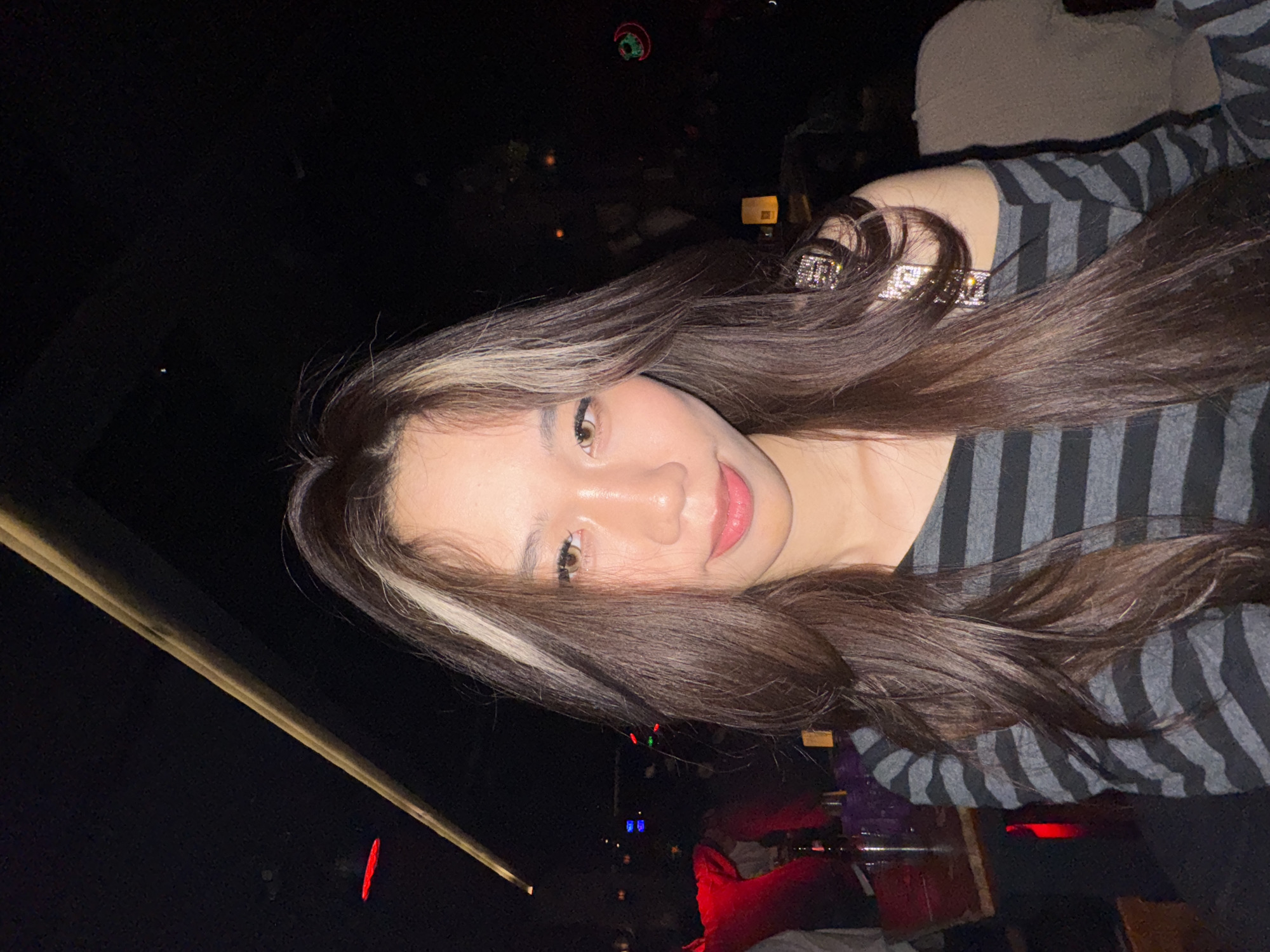 The image displays an East Asian female subject in an indoor establishment located in Liangjiang, China. The subject is positioned centrally, facing directly forward with a subtle, closed-mouth smile. She has long, dark brown hair with prominent blonde streaks framing her face, styled to fall over her shoulders. Her eyes are light brown, and she has visible eye makeup. She is wearing a long-sleeved, off-the-shoulder top with horizontal black and grey stripes; a reflective, possibly bejeweled strap or detail is present on her right shoulder. The environment is dimly lit, with a dark ceiling featuring a diagonal linear light fixture in the upper left and a faint red horizontal light further to the left. In the blurred background, various colored lights are visible, including red, green, blue, an orange glowing lantern on the right, and a circular green neon-like design on the far right. A wooden table with dark bottles is partially visible on the left, alongside a blurred individual in a red garment. An indistinct light brown upholstered object or person is present on the far right. No specific actions or interactions beyond the subject's direct gaze at the camera are discernible.