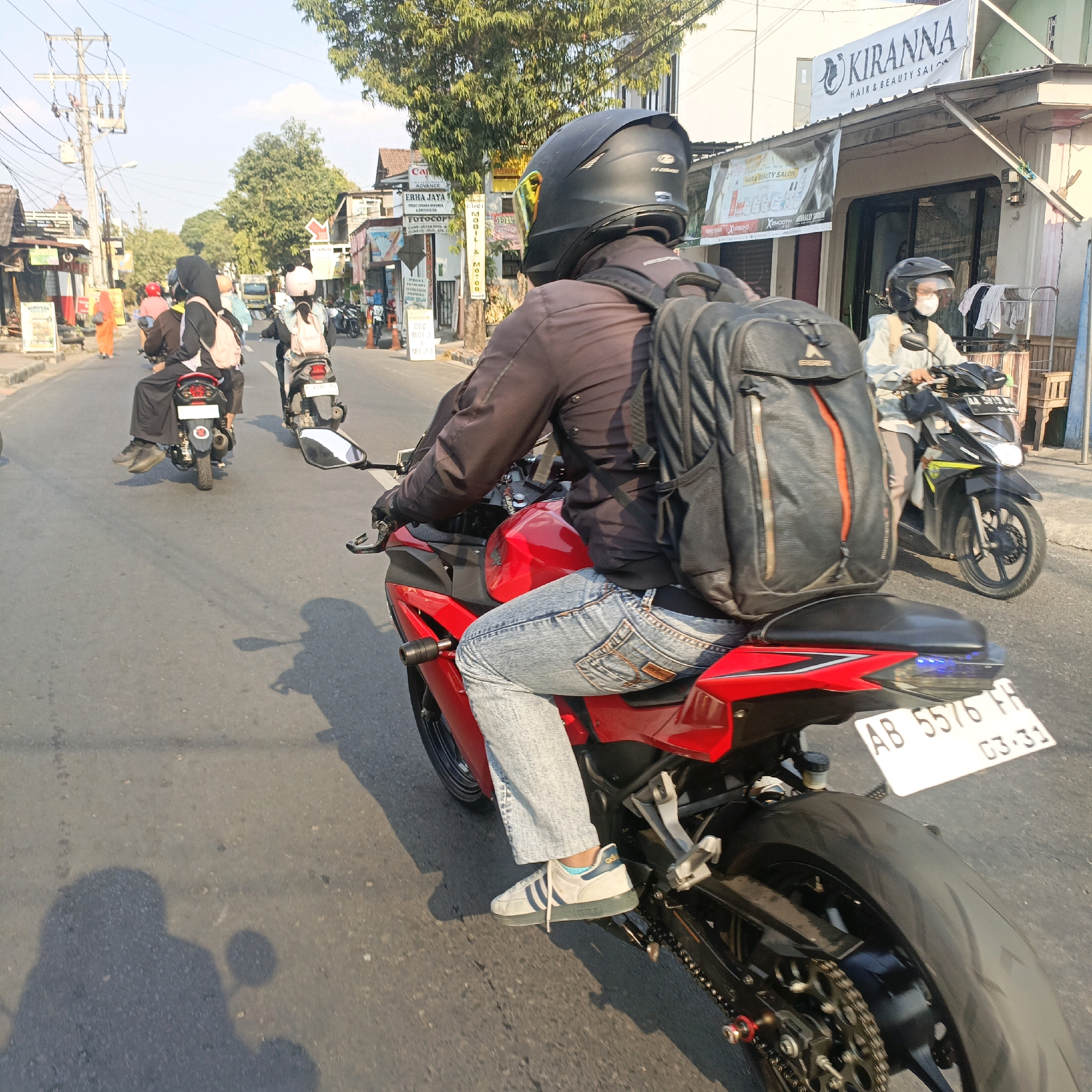 The image captures a street scene in Salatiga, Indonesia, with a focus on motorcycles and their riders. In the foreground, a person on a red motorcycle, seen from behind, is wearing a black helmet, a dark jacket, and light blue jeans. They are also carrying a black backpack with an orange accent. Their white sneakers with blue stripes are visible. To the right of the foreground motorcycle, another motorcycle is partially visible, with a rider wearing a helmet and a light-colored jacket. Further down the street, more motorcycles and riders are present.
The environment is a paved road lined with buildings and trees. Signs on the buildings indicate businesses, including "KIRANNA HAIR & BEAUTY SALON". The sky is bright and clear, suggesting daytime. The overall impression is of a busy urban street with typical Indonesian traffic. The license plate on the red motorcycle is partially visible and reads "AB 5576 FR 03-31".