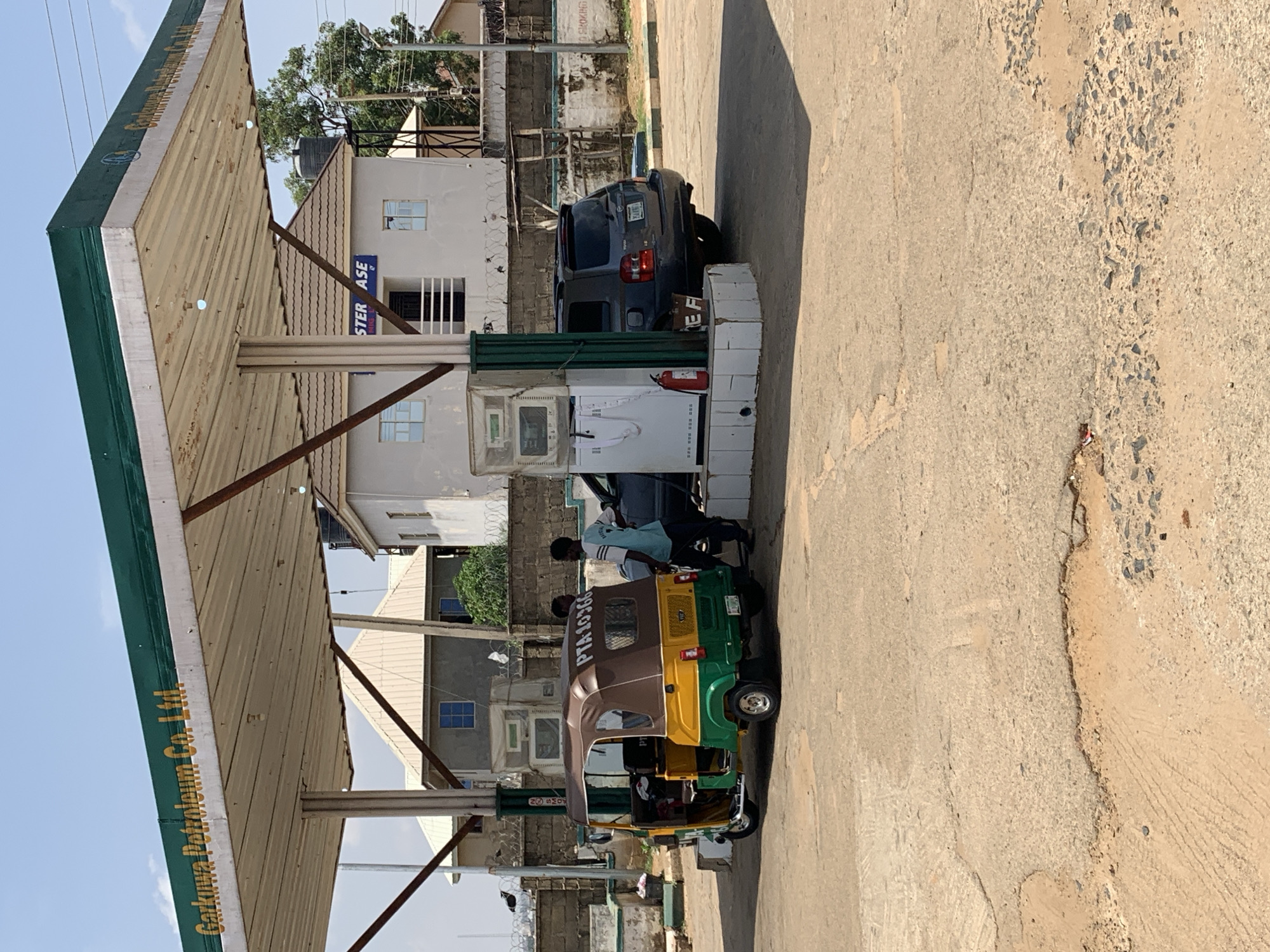 The image shows a gas station in Rafin Par, Nigeria. The gas station, identified by the sign "Gaskiya Petroleum Co. Ltd", features a typical forecourt canopy supported by metal pillars. A dark-colored SUV is parked at one of the fuel pumps. A three-wheeled taxi, known as a "Keke Napep," is also at the pump being refueled. There is a man wearing a light blue shirt visible behind the vehicle, presumably refueling it. Buildings line the street in the background, including a structure with a sign labeled "STER ASE." The road is made of asphalt and is worn in several places. The scene is bathed in sunlight.