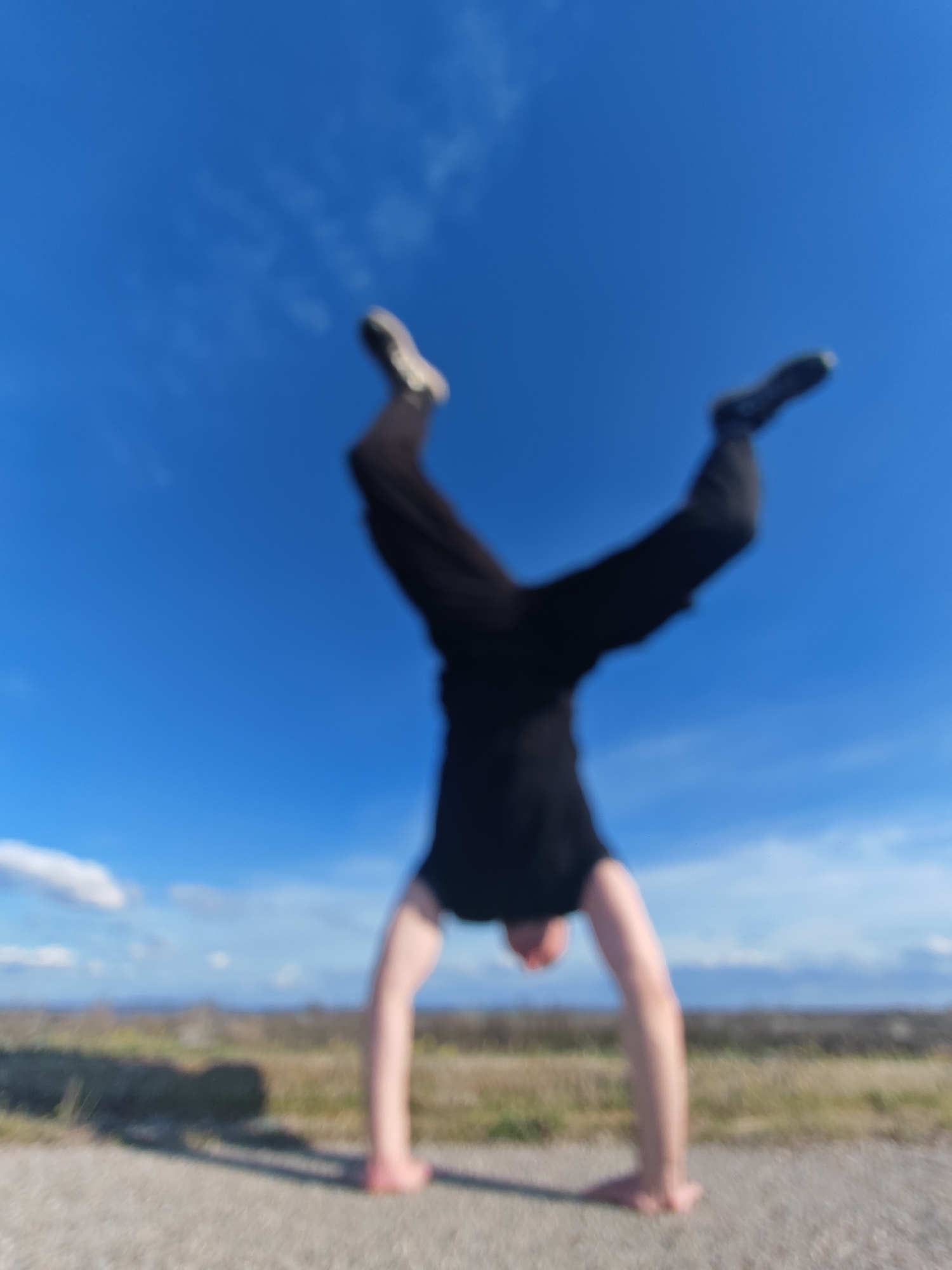 A blurred image shows a person doing a handstand outdoors on a paved surface with a grassy field in the background and a clear blue sky with light clouds above. The person is wearing dark clothing, including pants and a shirt, and appears to have sneakers on their feet. The location seems to be an open area in Shumen, Bulgaria, based on the landscape and environment, although no specific landmarks are visible due to the blurriness. The primary action is the handstand being performed by the individual.