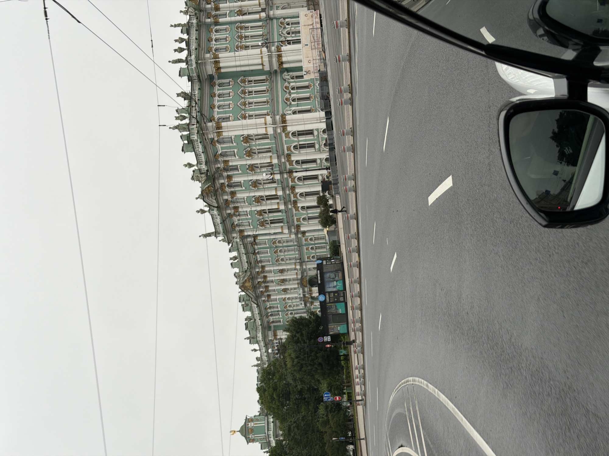 The image captures a view of the Winter Palace, a landmark building in Saint Petersburg, Russia, from a moving vehicle. The palace, painted in a distinctive pale green with white and gold trim, dominates the background. Its facade features numerous windows and decorative sculptures along the roofline. In the foreground, a wet asphalt road curves to the left, marked with white lane lines. The sky is overcast and gray. The reflection in the car's side mirror shows trees and buildings. A small kiosk with an "INFORMATION" sign is visible on the sidewalk in front of the palace, and a traffic light with a red signal is also present. Several concrete bollards line the edge of the road, separating it from a pedestrian area.