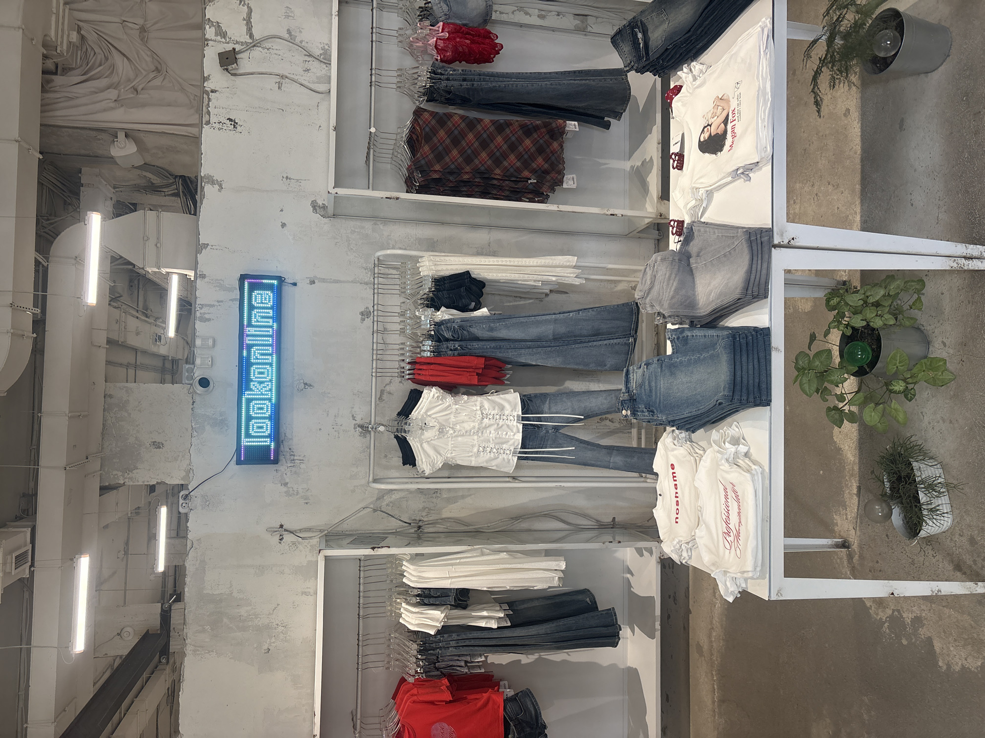 This is an interior shot of a retail store, likely in Moscow, Russia, indicated by the Cyrillic text on the vertical digital sign that reads "lookinlab." The sign is mounted on a distressed white wall. The store appears to be selling clothing, as multiple racks are visible, stocked with items like t-shirts, jeans, and plaid garments. The clothes are hung on metal racks, and the style of the clothes suggests a trendy, modern aesthetic.
Next to the racks, there is a metal display stand. The topmost level displays a stack of t-shirts with a graphic print of a woman and the name "Megan Fox" alongside a pile of folded denim. The middle rack contains stacked jeans and t-shirts. The bottom rack has multiple white t-shirts with the word “noshame” and another word in a cursive font.
Next to the display stand on the right is a small potted plant with green foliage. On the top is another plant in a metallic pot.
The overall lighting is bright and industrial, enhanced by fluorescent tube lighting on the ceiling, which contributes to the stark, urban aesthetic. The walls and ceiling display a raw, unfinished look with exposed piping and concrete.