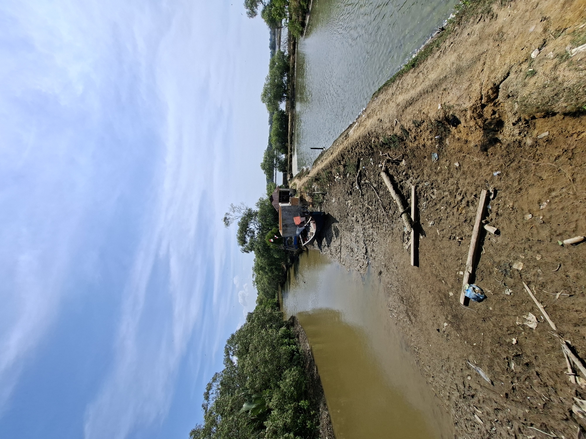 The image depicts a tranquil waterfront scene in Kruengmate, Indonesia. A narrow, muddy river, bordered by lush green trees, flows into a wider body of water on the right. The riverbank is characterized by exposed mud and scattered debris, including pieces of wood and plastic waste. A small, weathered boat rests on the bank near a simple, tin-roofed structure. The sky above is a bright blue, dotted with wispy white clouds. The overall impression is one of a rural, somewhat undeveloped coastal area.