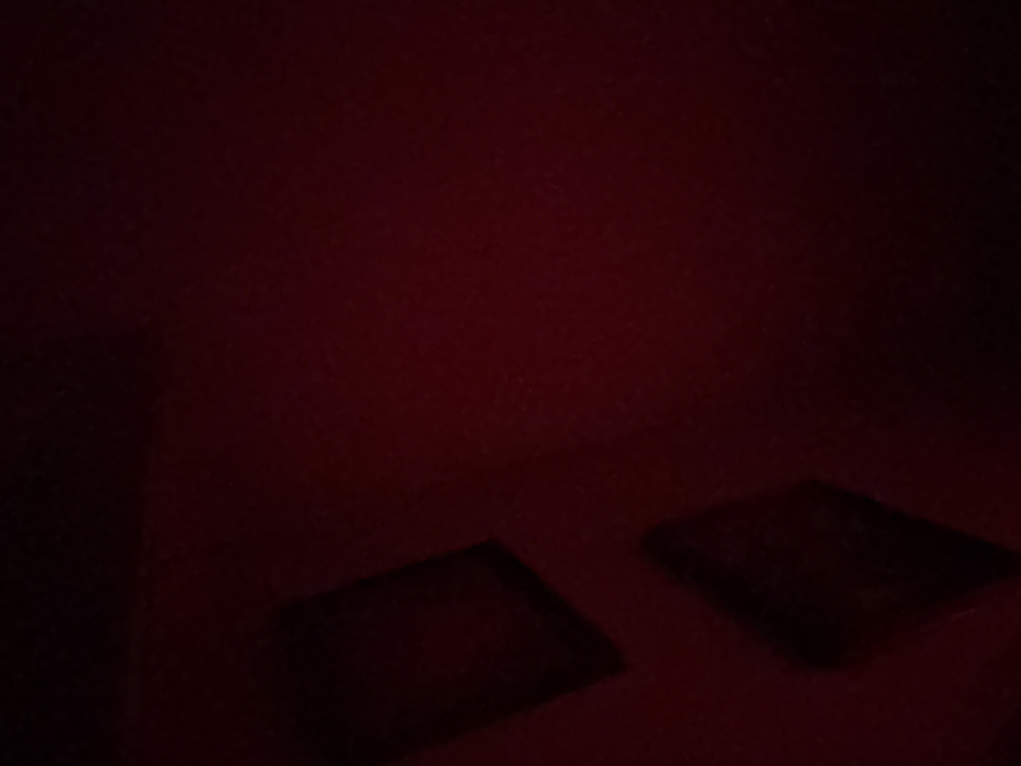The image depicts a dimly lit interior space with a red-toned palette. Two dark, rectangular objects are situated on a surface in the lower portion of the frame. The left object is more pronounced and appears closer, with a distinct edge suggesting a three-dimensional form. The other object is slightly more distant and less defined. The environment is generally dark, with the objects appearing as silhouettes against a red-hued background. The absence of discernible details prevents further identification of the objects or the space's exact nature. There is no information available to determine whether this is Troyan, Bulgaria.