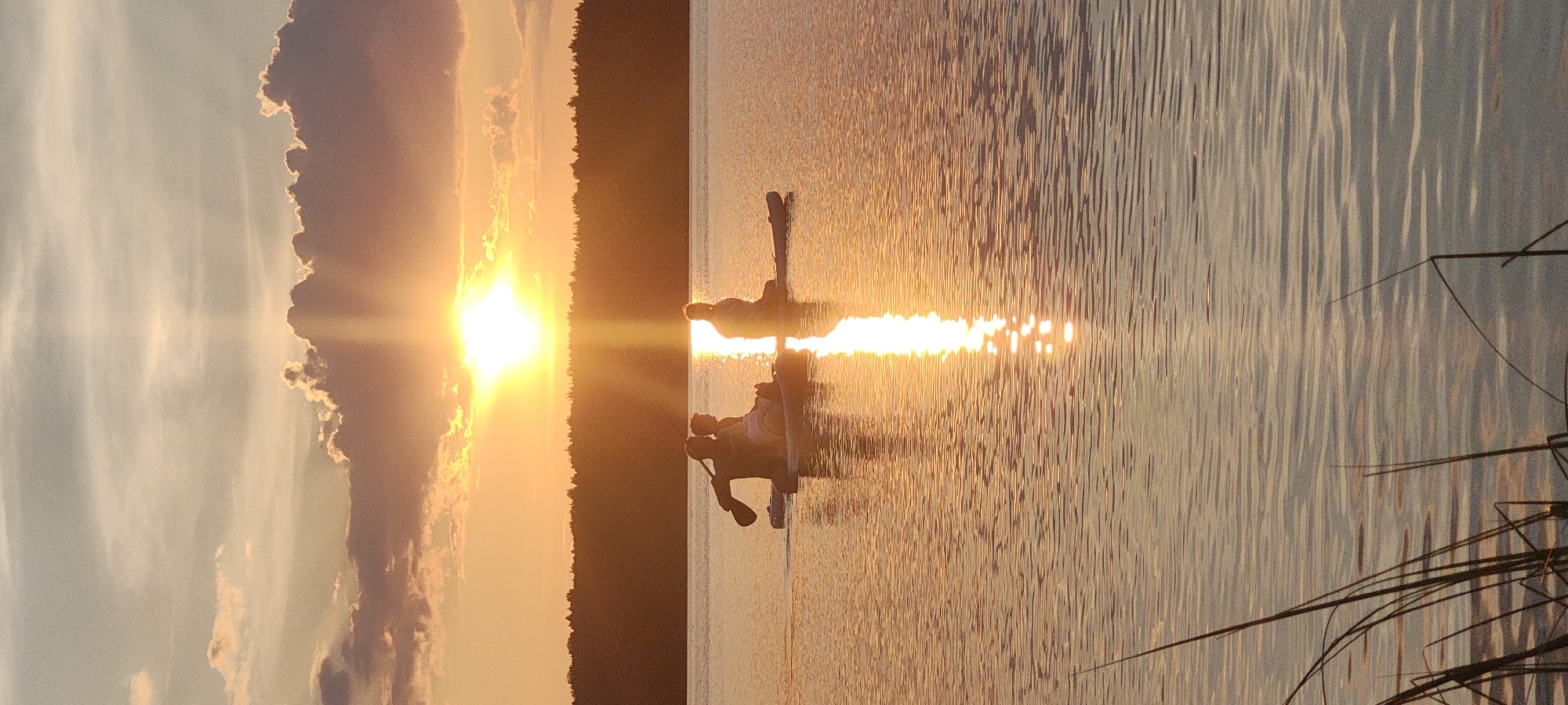 The image depicts a serene sunset over Mednoye Ozero, Russia. The sky is a canvas of warm hues, with the sun a bright orb just above the horizon, casting a golden reflection on the rippling water. Three individuals are seen on paddleboards, silhouetted against the sunset. One person in the foreground is actively paddling, while the other two appear to be seated. The background features a dense line of trees, forming a dark silhouette against the vibrant sky. The foreground shows the edge of the lake with tall grass. The overall scene conveys a sense of tranquility and peacefulness.