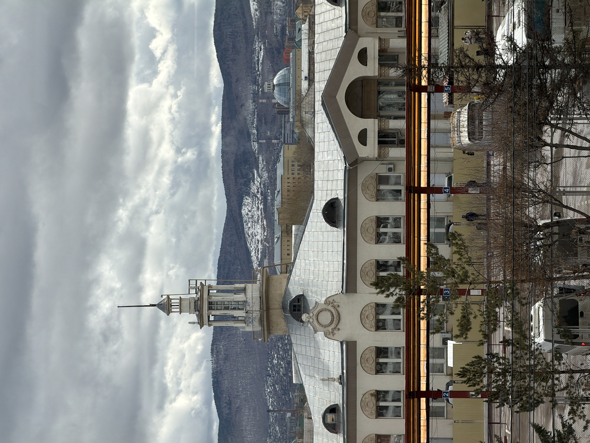 The image shows a building in Krasnoyarsk, Russia. The building has a tower with a spire on top. The building also has a series of arched windows. Behind the building are mountains. In front of the building are trees and a street with vehicles. There are people on the street. The sky is cloudy.