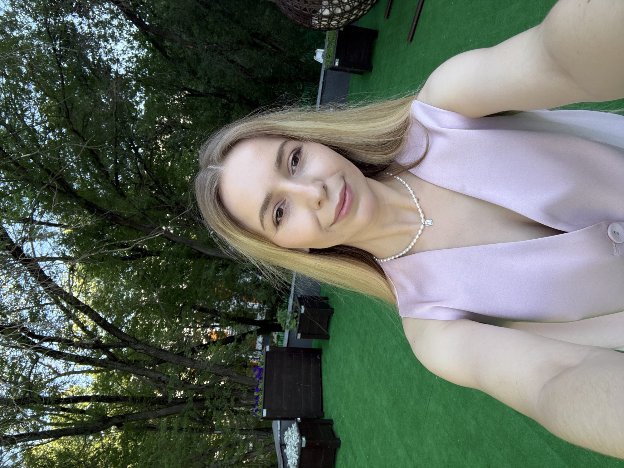 A woman with blonde hair and fair skin, wearing a light pink V-neck top and a pearl necklace with a diamond pendant, takes a selfie outdoors. She has light makeup and is smiling. The background features lush green trees and a bright blue sky. In the foreground, there is a bright green carpeted area. Several dark wooden planters are visible, some containing purple flowers and white decorative stones. A woven hanging chair and other outdoor furniture can be seen in the distance. The overall scene suggests a relaxed outdoor setting, potentially a rooftop terrace or a garden.