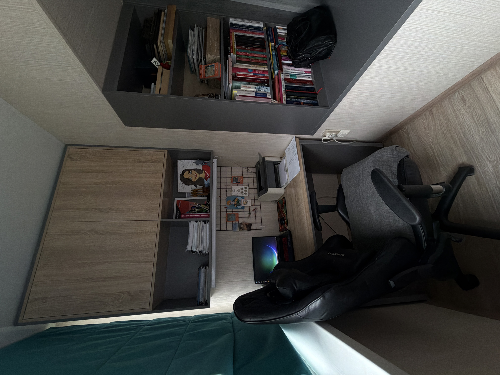 The image shows an interior view of a room. The scene includes a bed with blue bedding, a built-in storage unit with wooden-colored cabinets and shelves, a desk area, a gray office chair, a monitor, and books. The room is filled with objects that suggest a personal living or study space. There is no indication of the location within Birobidzhan, Russia, or any specifics about any people.