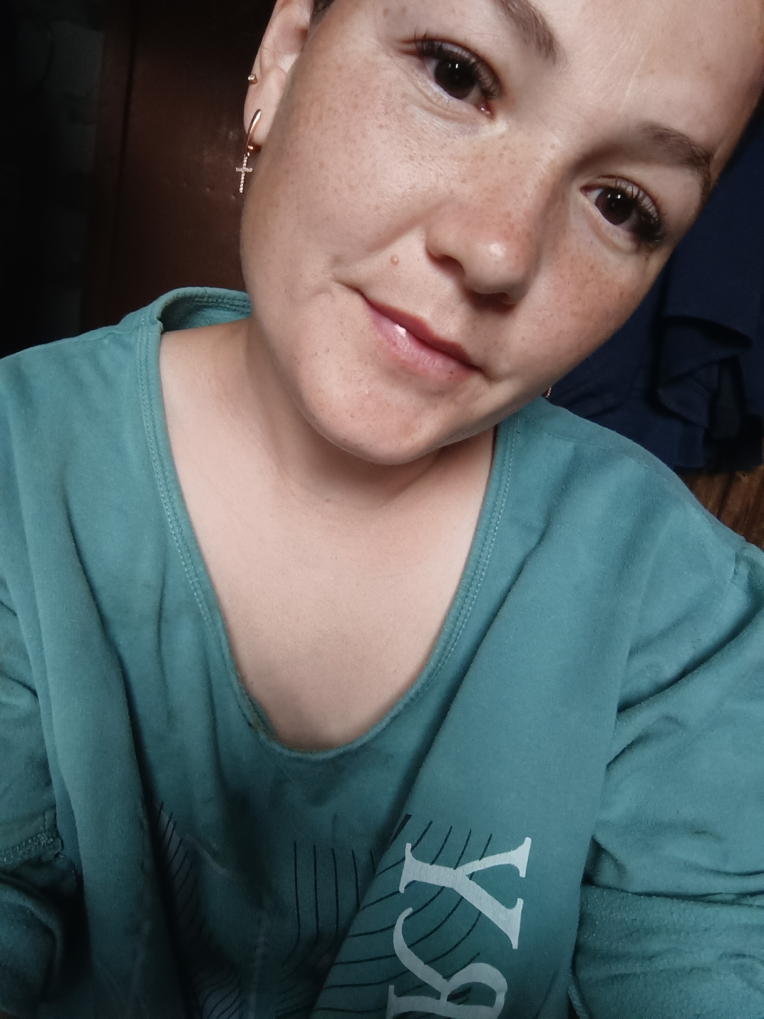A close-up, medium-angle shot captures the face and upper body of a young woman wearing a teal-colored t-shirt. The woman has fair skin with freckles, dark brown eyes, and light brown eyebrows. She has on mascara and possibly eyeliner, and her lips are lightly colored. A cross-shaped earring is visible in her left ear. The t-shirt has a V-neck and a graphic print on the right side of the chest. The background is out of focus, with a dark brown wall or door on the left and a dark blue garment on the right. The lighting suggests it is daytime, and the overall impression is of an indoor, casual setting. The image is a selfie taken by the woman herself.