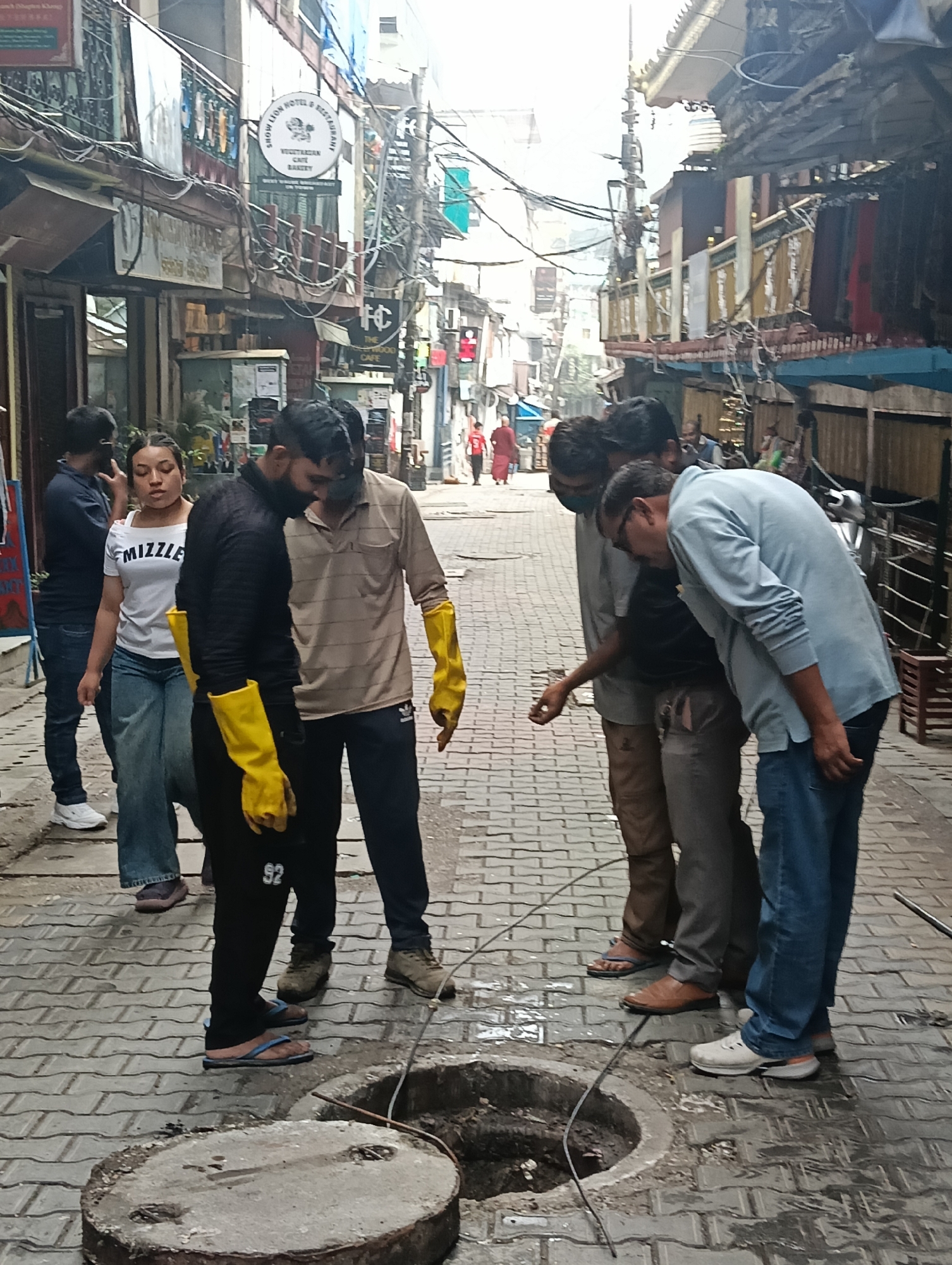 In Bhagsu, India, a group of individuals are gathered around an open manhole on a cobblestone street. A young woman, wearing a white off-the-shoulder top with "MIZZLE" printed on it and blue jeans, stands to the left. To her right, a man wearing a black long-sleeved shirt and black pants with yellow rubber gloves is looking down into the manhole. Another man, wearing a light brown striped shirt and dark pants with yellow rubber gloves, stands next to him, also looking down. Two other men are visible in the background, one in a light blue shirt and jeans, and another in a gray shirt. The street is lined with buildings featuring signs for shops and businesses, including "SHOW LION HOTEL & RESTAURANT" and "THE WOOD CAFE". Overhead, a dense network of electrical wires crisscrosses the sky. The ground is wet, indicating recent rain or cleaning.