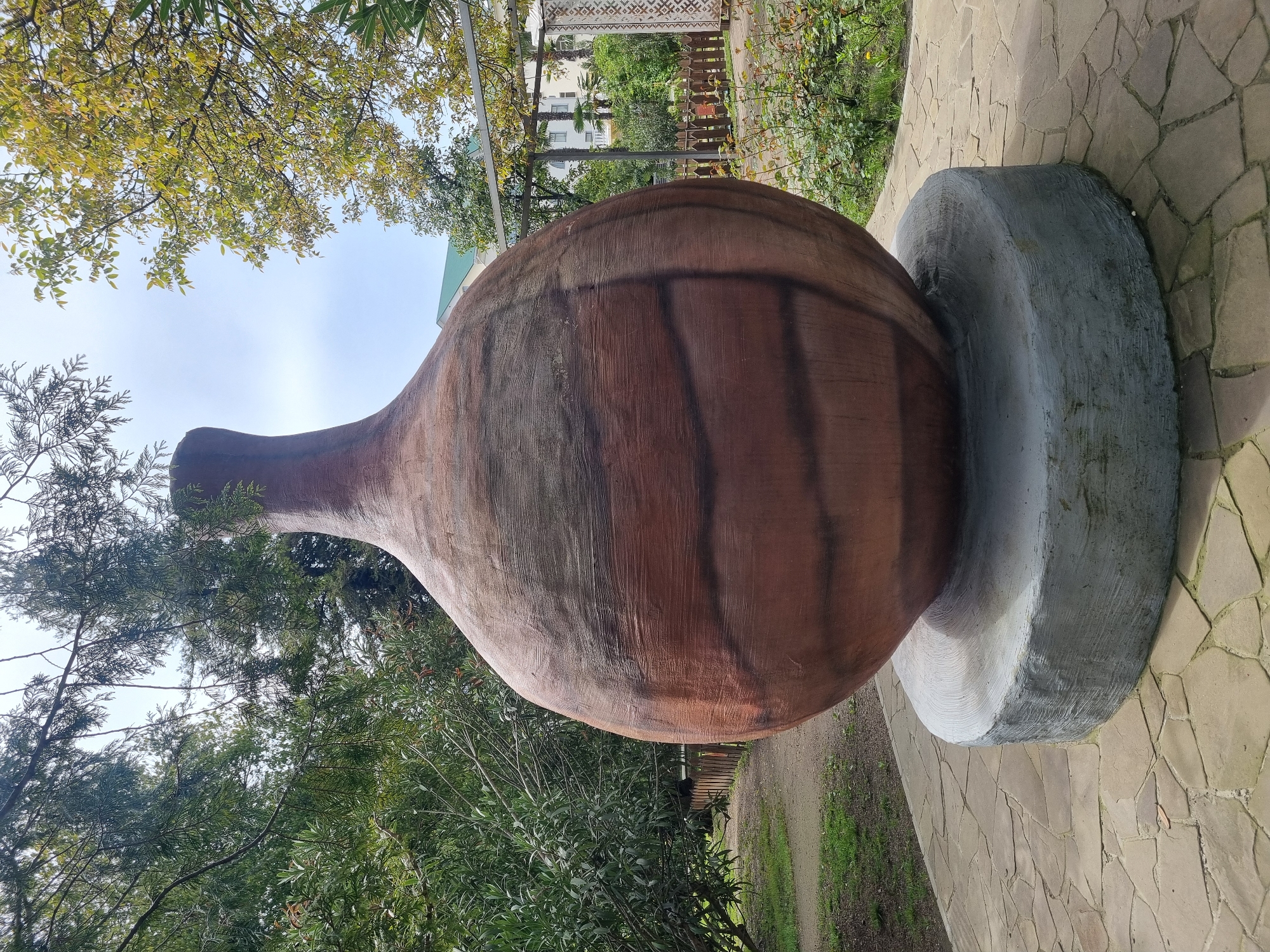 A large, brown, vaguely amphora-shaped sculpture is prominently displayed in a garden-like setting in Sochi, Russia. The sculpture appears to be made of concrete or a similar material, painted to resemble an earthenware vessel. It rests on a round, gray concrete base. The ground is paved with irregular stone slabs. The surrounding vegetation includes trees and shrubs. In the background, there are structures, possibly buildings, obscured by the foliage. The sky is visible through the trees.