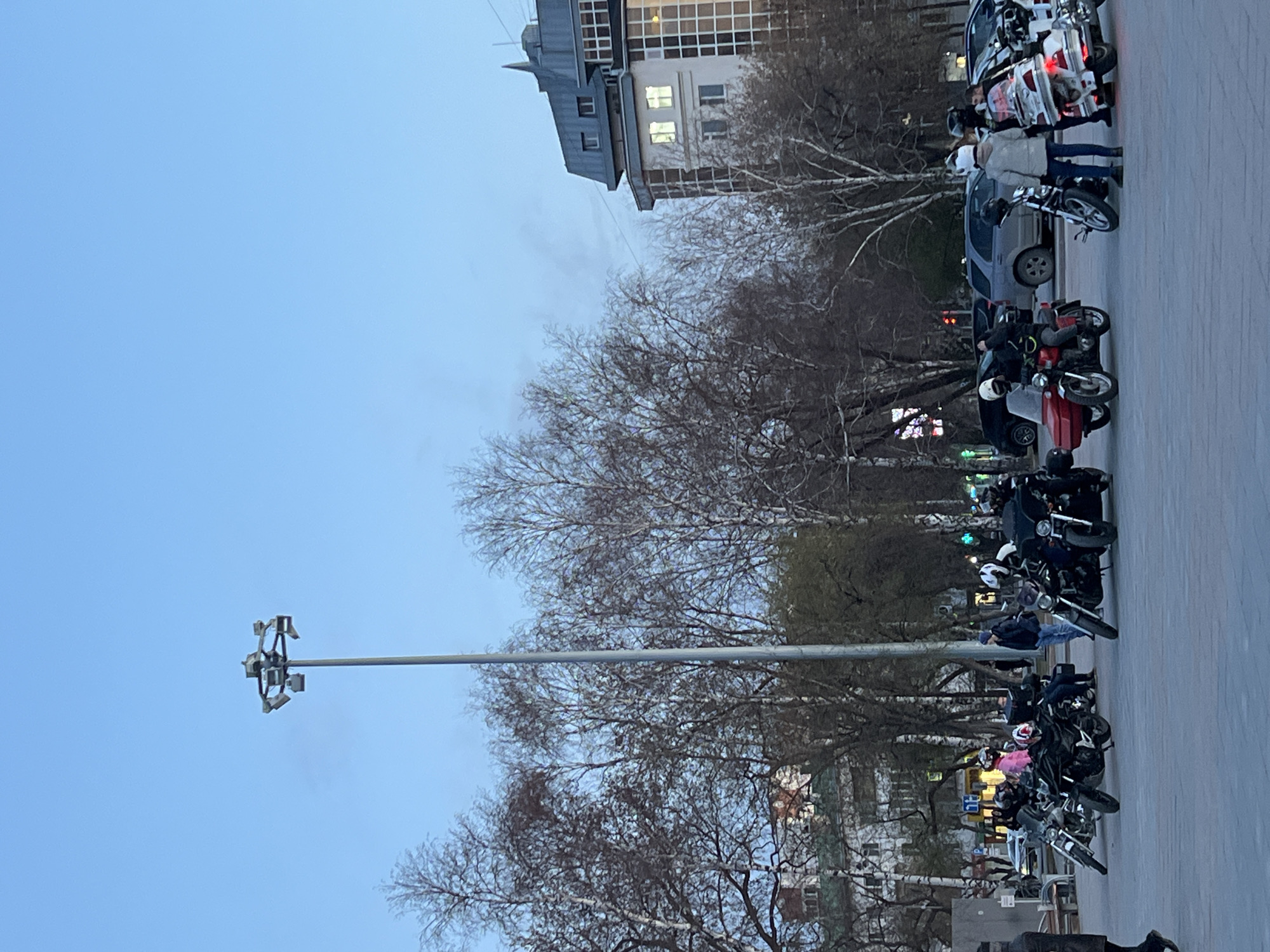 The image shows a street in Tyumen, Russia. Several motorcycles are parked in a row on the right side of the frame, with a few people standing and sitting nearby. Trees with bare branches are scattered throughout the scene. A tall streetlight is visible, reaching into the blue sky. In the background, there is a building with several windows. The overall lighting suggests it is either early morning or late evening.