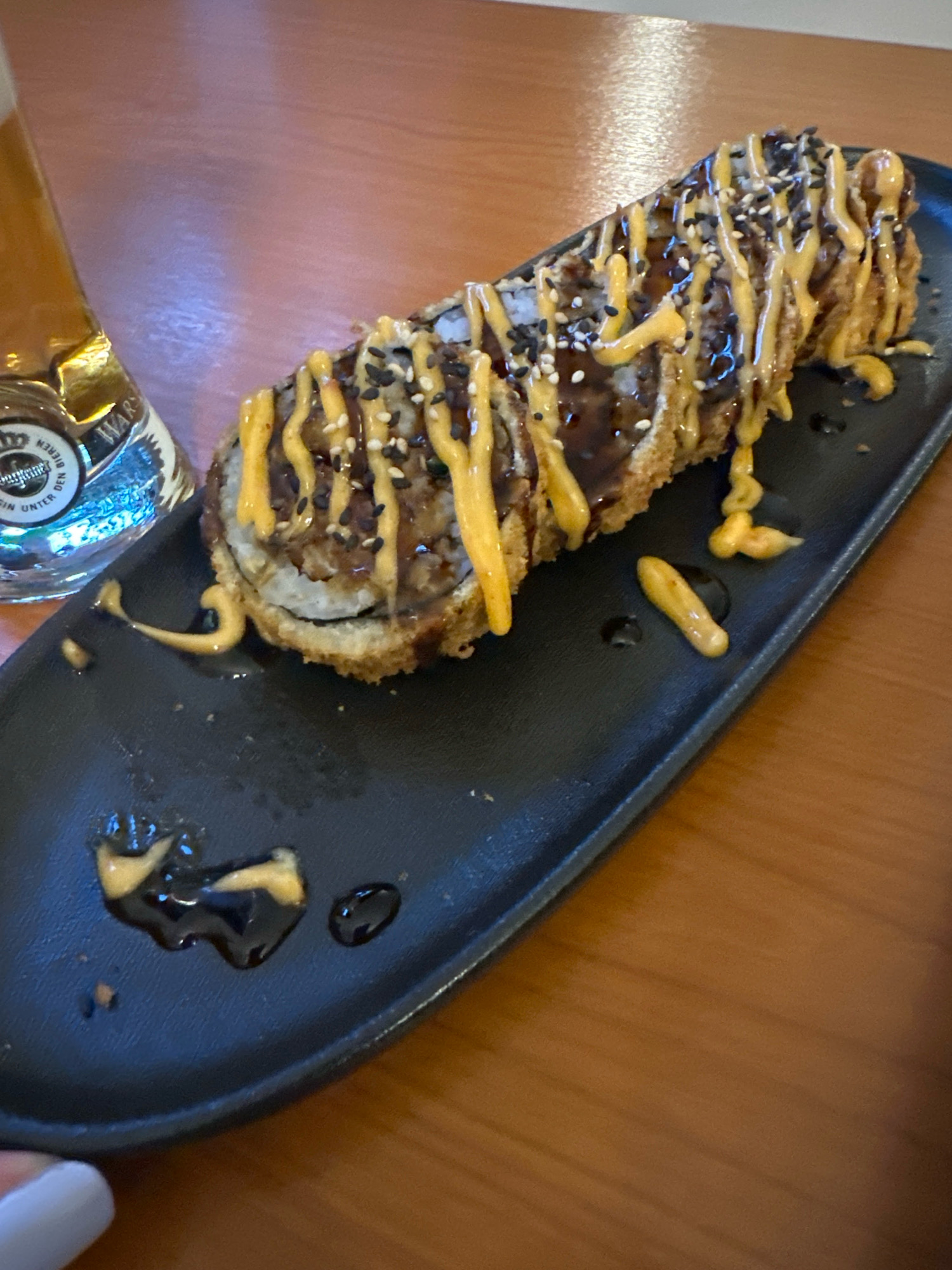 A single, deep-fried sushi roll, drizzled with dark sauce and a yellow sauce, sits on a black rectangular plate. The sushi is topped with sesame seeds. A glass of beer, partially visible on the left side of the frame, contains liquid and has a label with text that appears to read "WARSCHAUER BIEREN" and "UNTER DEN..." The scene is set on a brown wooden table, suggesting an indoor dining setting, possibly a restaurant or bar in Krefeld, Germany. There are also small droplets of the sauces on the plate, as well as a fingerprint smudge on the bottom left corner of the plate.