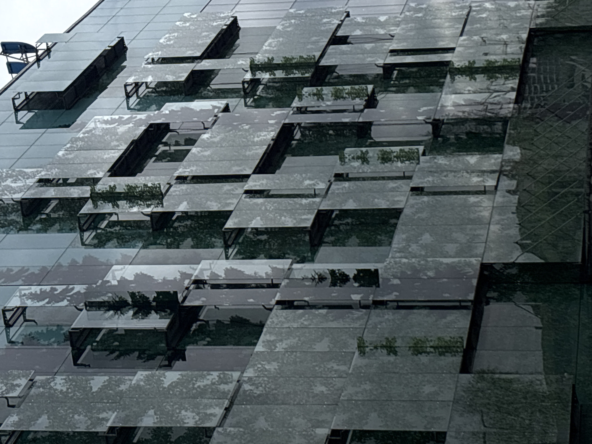 The image shows a close-up of a modern building facade in Moscow, Russia. The facade features a pattern of irregularly staggered, cantilevered balconies or ledges, covered with reflective material that reflects the surrounding environment, including trees. There is also a work platform affixed to the facade with a chair. The overall tone is monochromatic, with shades of gray and green predominating.