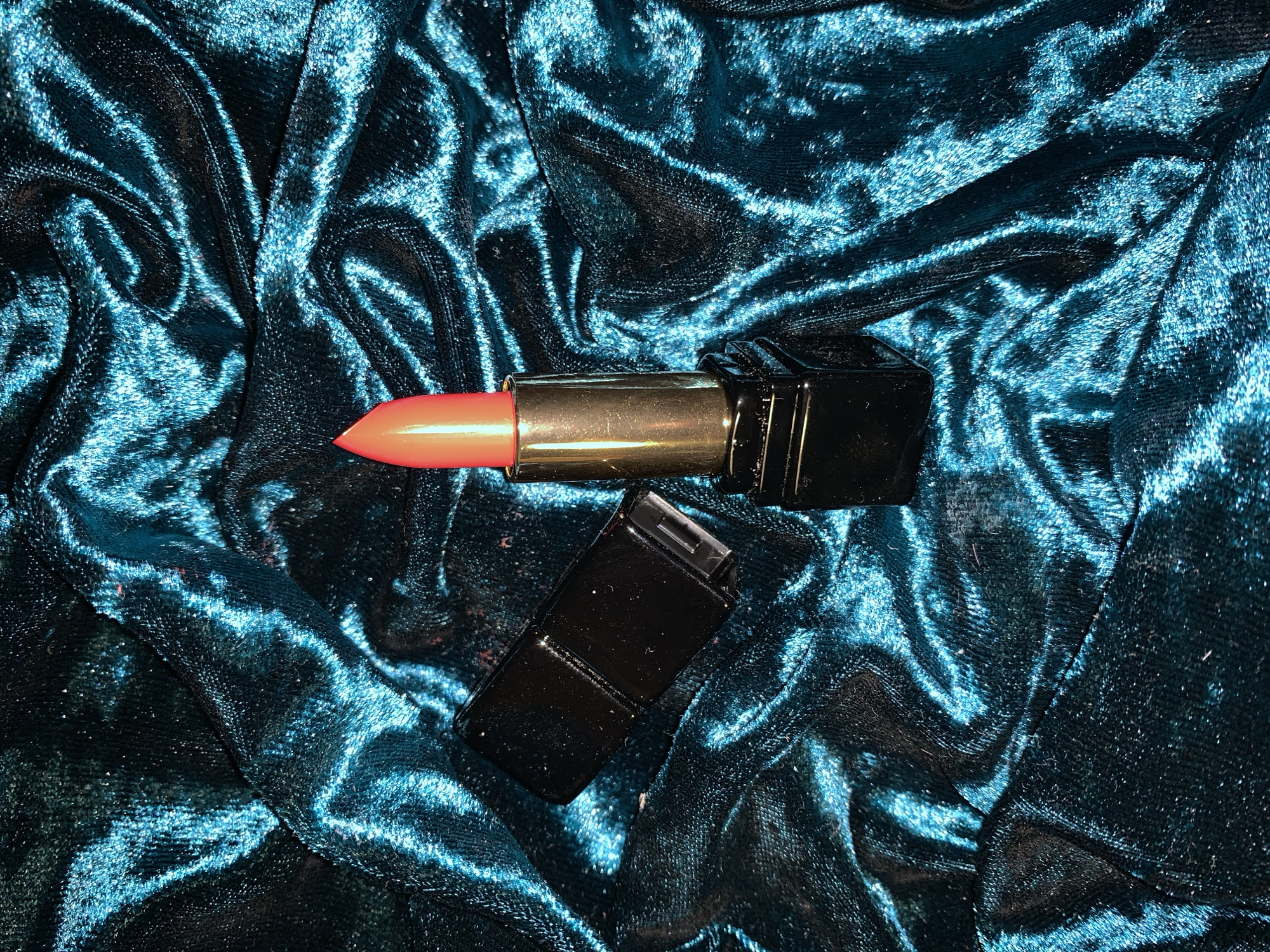 The image shows an opened lipstick tube with a coral color lipstick lying on a crumpled, dark blue velvet fabric. The lipstick tube has a golden middle section and a black casing. The lid of the lipstick is beside the opened tube. The background consists of the textured surface of the velvet fabric. The location and presence of people are unknown.