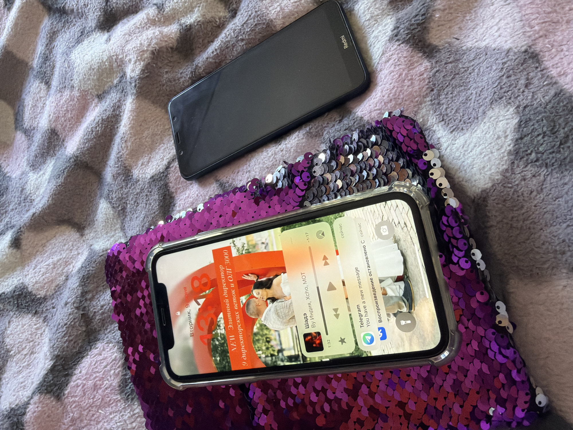 In Raevka, Russia, two smartphones are placed on a textured, pink and gray checkered blanket. The phone on the left is an iPhone with a clear case, displaying a lock screen. The screen shows the time "13:18" and the date "Вторник, 9 июня" (Tuesday, June 9). Below the time is text that reads "NEW Элитный директор 9 директорских веток и ОЛГ 3000" (NEW Elite Director 9 director branches and OLG 3000). A picture of a couple kissing is displayed, overlaid with music playback controls showing a song titled "Шадэ" by Индия, Xcho, MOT, with a duration of 1:17 and playback at 1:32. A Telegram notification states "You have a new message." The phone on the right is a black Redmi smartphone.