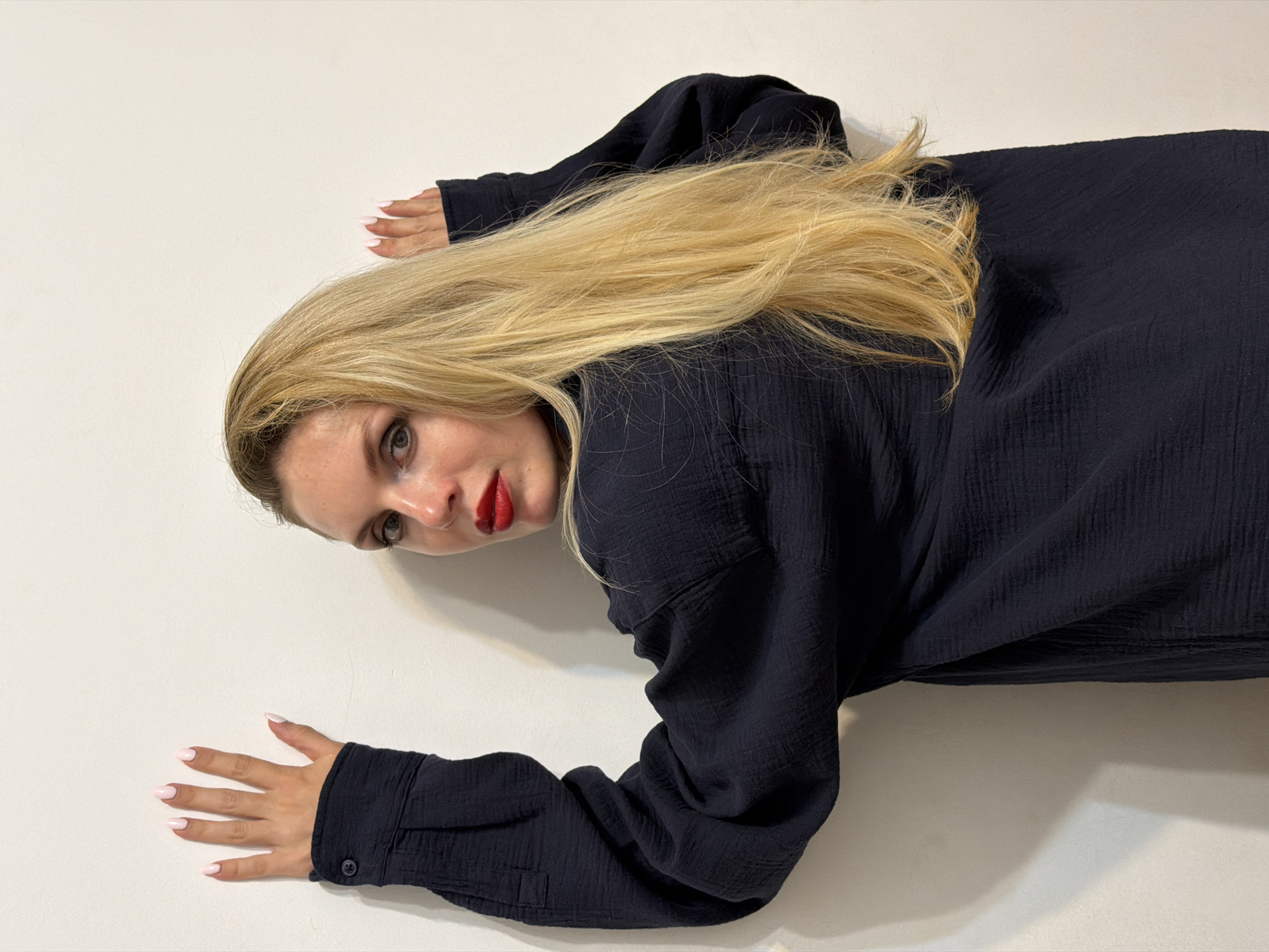 A woman with long blonde hair and red lipstick is lying on a white surface. She is wearing a dark blue shirt with long sleeves. Her nails are painted a light pink. The scene is indoors and the background is plain white. The location is not apparent.