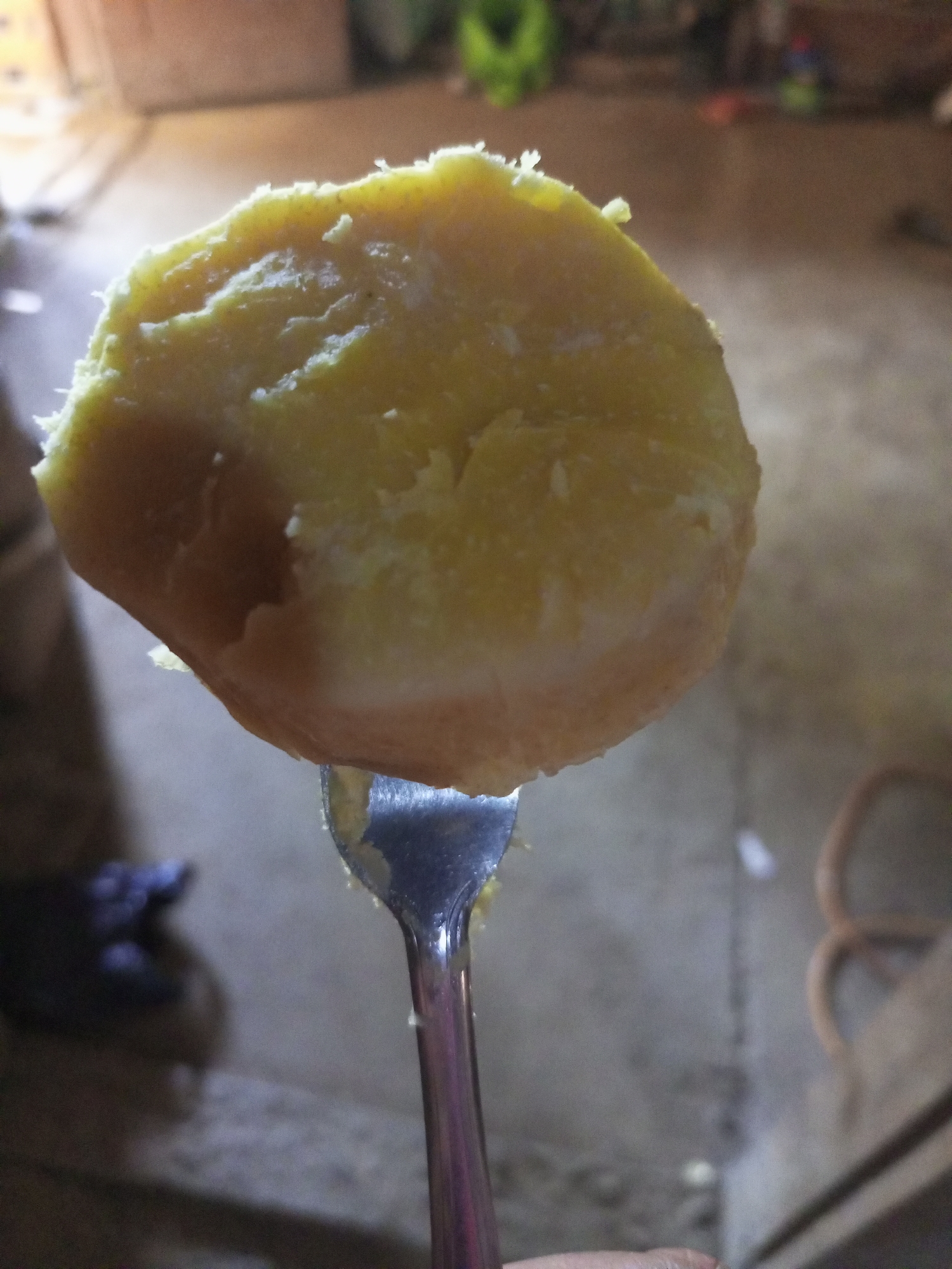 A close-up, shallow-depth-of-field shot shows a slice of cooked sweet potato speared on a metal fork, held up in the foreground. The sweet potato is a light yellow-orange, with a rough, slightly textured surface and some visible white fibers at the edges. The fork's tines are partially embedded in the potato. The background is blurred, suggesting an indoor environment with a concrete or dirt floor and what appears to be a wooden or brown wall in the upper portion. Hints of green and red objects are visible in the far background, possibly in a doorway or on a shelf. The lighting is dim and warm, casting soft shadows. This image was taken in Jatipurno, Indonesia.