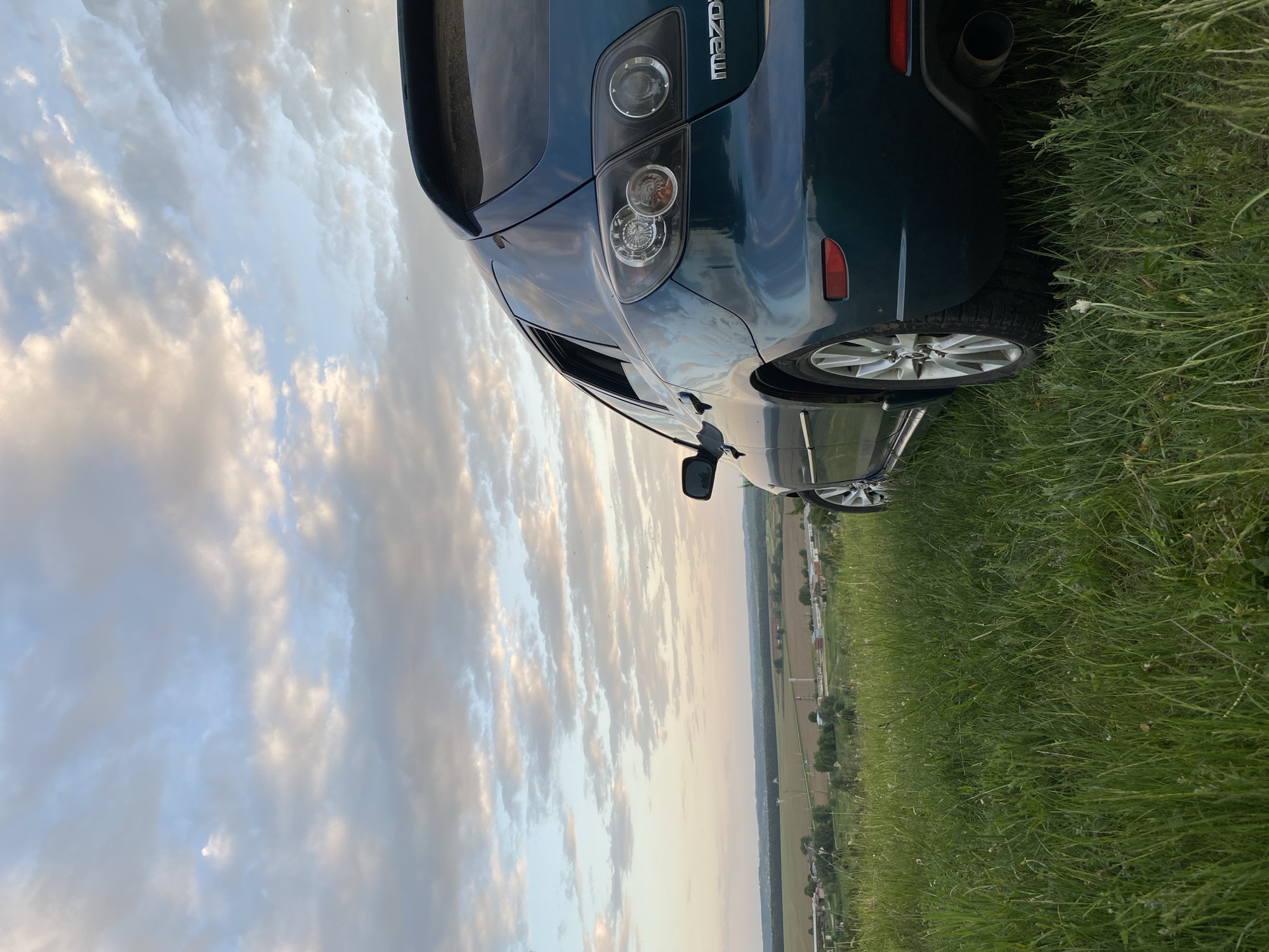 The image depicts a blue Mazda 3 parked on a grassy field in Unknown, Unknown. The rear of the car is visible, including the taillights, exhaust pipe, and rear tire. The car is partially surrounded by tall grass. In the background, a landscape is visible, including fields, trees, and buildings, under a sky filled with clouds.