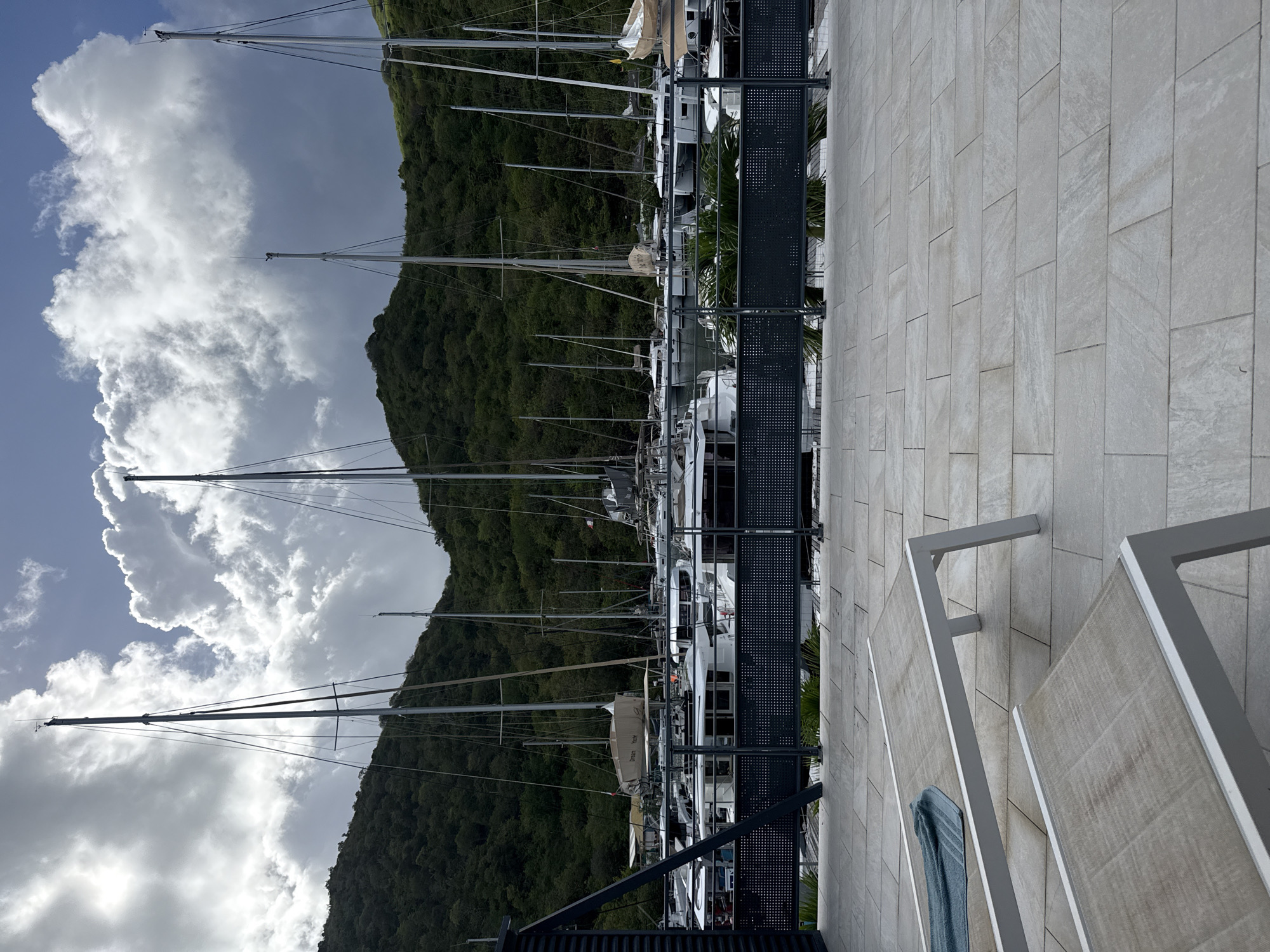 The image depicts a marina in Saint-Martin, France, with numerous sailboats docked. Tall masts rise against a backdrop of a lush green hillside and a sky filled with dramatic, billowing clouds. The marina's architecture includes a light-colored, tiled walkway with modern railings and sunshades. A dark metal fence separates the walkway from the moored boats. A blue towel is draped over the railing.