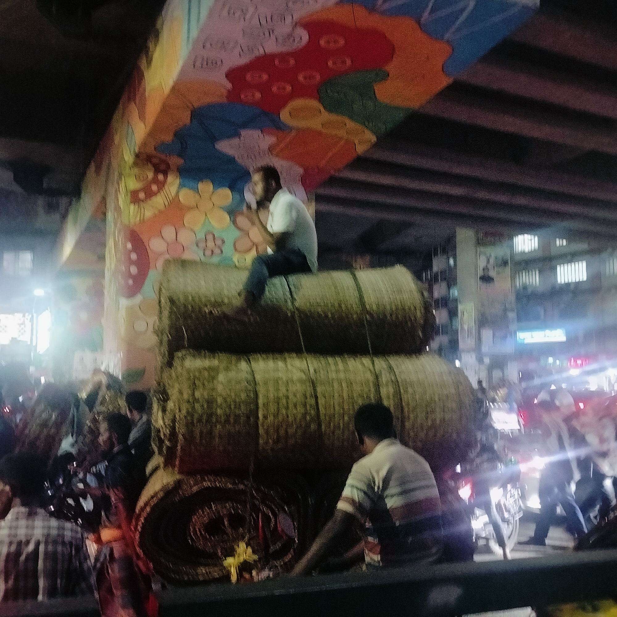 The image depicts a street scene in an urban environment, possibly at night. A large, painted support pillar is visible, with colorful designs of flowers and abstract shapes. In front of the pillar, a stack of bundled, cylindrical objects, possibly hay or straw, is present. A man is sitting atop the stacked objects, while another man stands near the base of the stack. The scene is crowded, with people and motorcycles visible in the background. The lighting is a mixture of artificial lights from street lamps and vehicle headlights. The exact location and time are not specified, but the image suggests a busy street in a city.