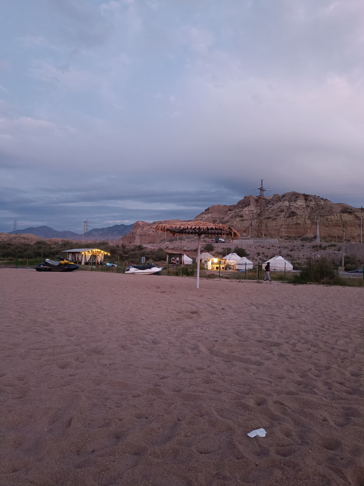 This image depicts a sandy beach area with several yurts and a covered structure in the background, set against a backdrop of hills and a cloudy sky. The scene appears to be at dusk or dawn.
In the foreground, the sand is textured with footprints and a piece of white litter. A thatched umbrella is visible on the left side of the frame, partially obscuring the view of some yurts.
In the mid-ground, a cluster of white, circular yurts are arranged, some illuminated from within. A person in a grey jacket and dark pants is walking away from the yurts towards the right. Adjacent to the yurts is a long, covered structure with white curtains, illuminated by warm lights, and what appears to be a small building behind it. Two jet skis are parked on the sand near this structure, one black and yellow, and the other white.
The background features rolling hills and rocky outcrops. A large electricity pylon stands prominently on the slope of one of the hills, with power lines extending across the landscape. The sky is a pale blue with scattered grey clouds, suggesting either early morning or late evening.
The scene is located in the vicinity of the city of Tong, Kyrgyzstan, likely at a lakeside or riverbank resort, given the presence of jet skis and the general landscape. The overall atmosphere is one of a quiet evening at a recreational area.