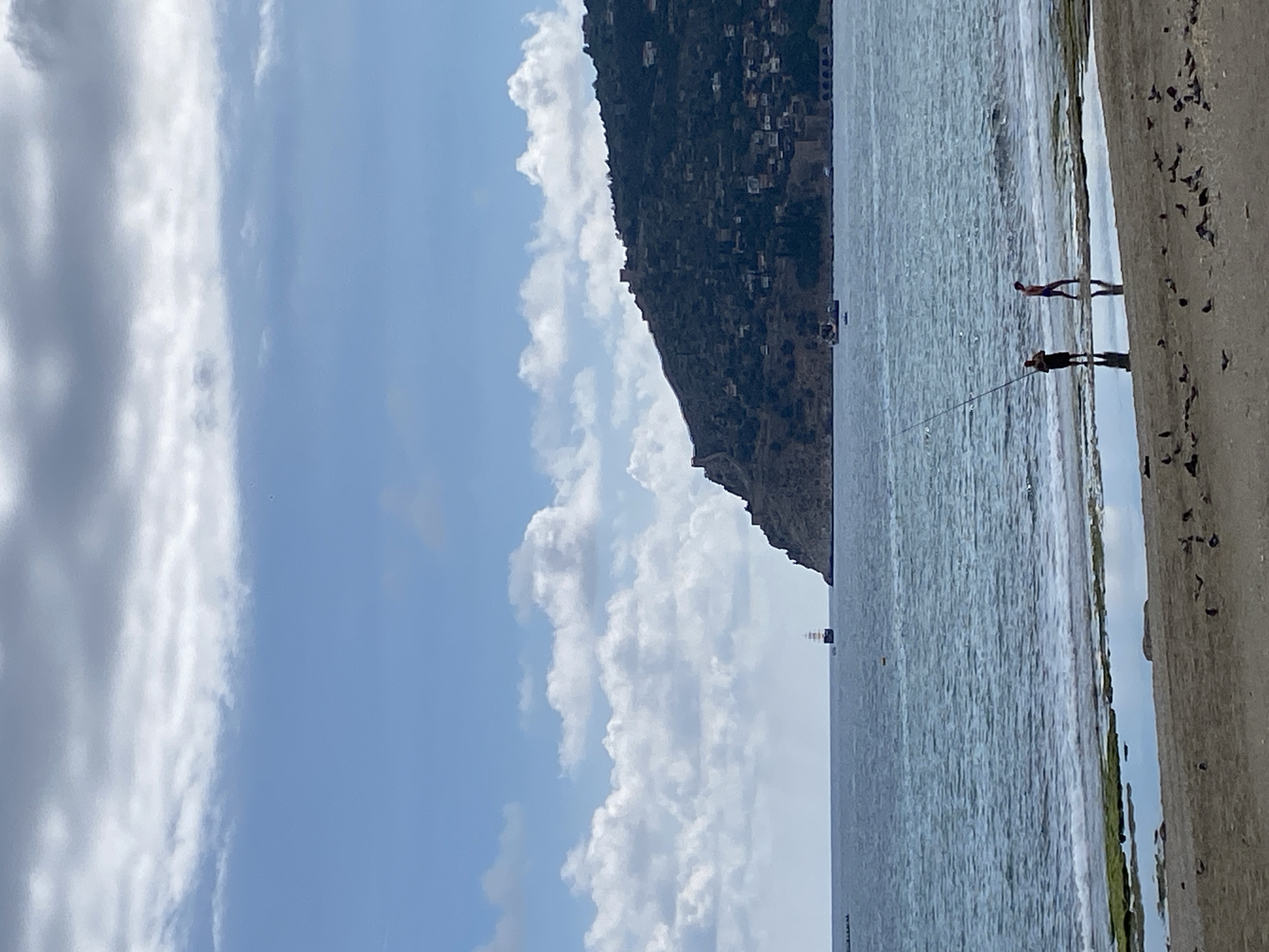 The image shows a coastal scene. On the right, a concrete shore is visible with a flock of birds perched on it. Two people stand near the water's edge, one appearing to be fishing with a rod. The ocean stretches out with small waves reaching the shoreline. A prominent mountain with clustered buildings on its slopes dominates the background. The sky is partly cloudy with a mix of blue sky and heavy clouds on the left. The setting suggests a location in Alanya, Turkey, given the coastal geography and building style on the mountain.