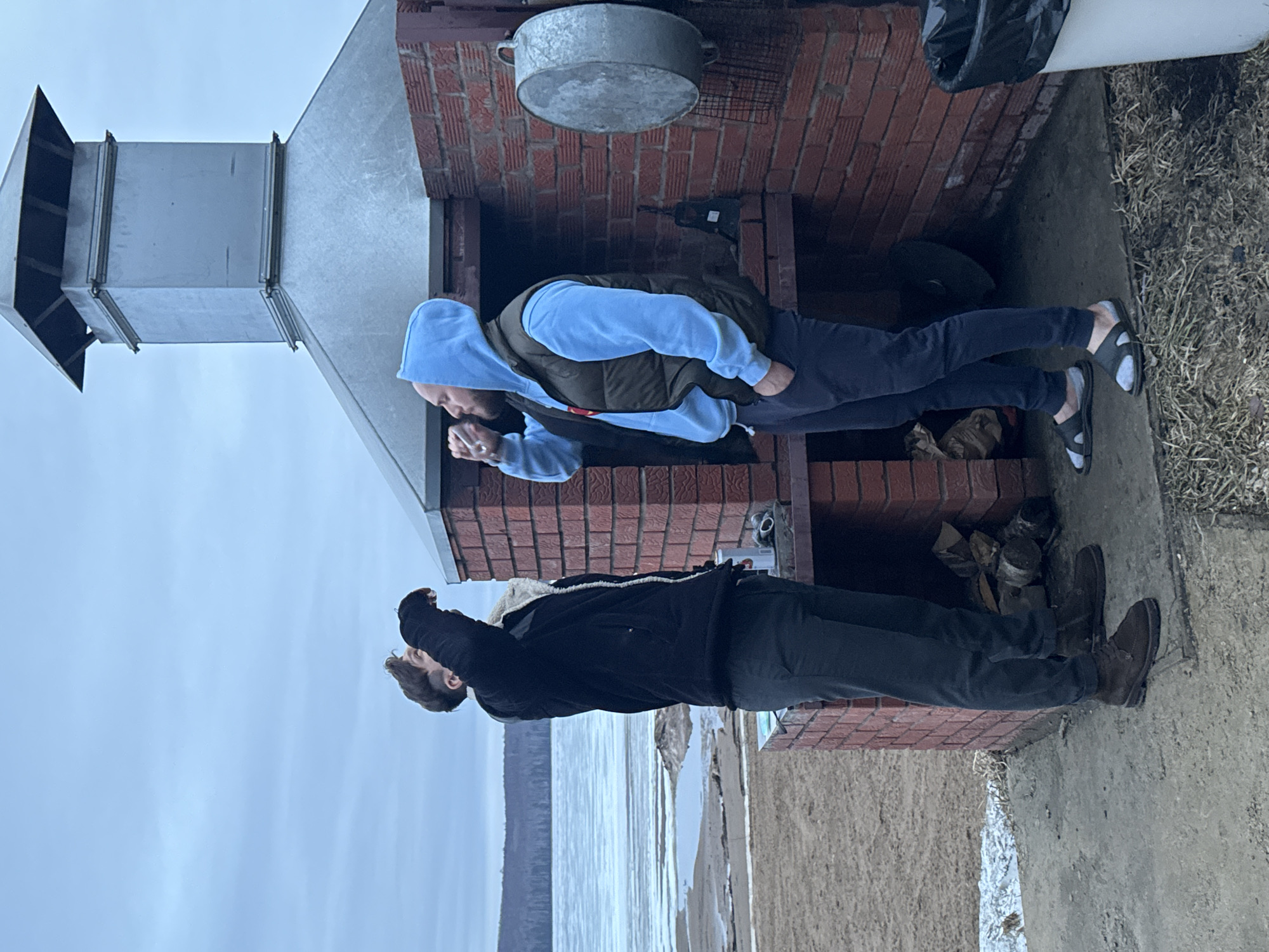 Two men stand in front of a brick outdoor fireplace with a metal chimney in Chaikovsky, Russia. The man on the left wears a black jacket and dark pants, while the man on the right wears a light blue hooded sweatshirt, a dark vest, and dark pants. The ground appears to be partially covered with snow. There is water and a shore visible in the background.