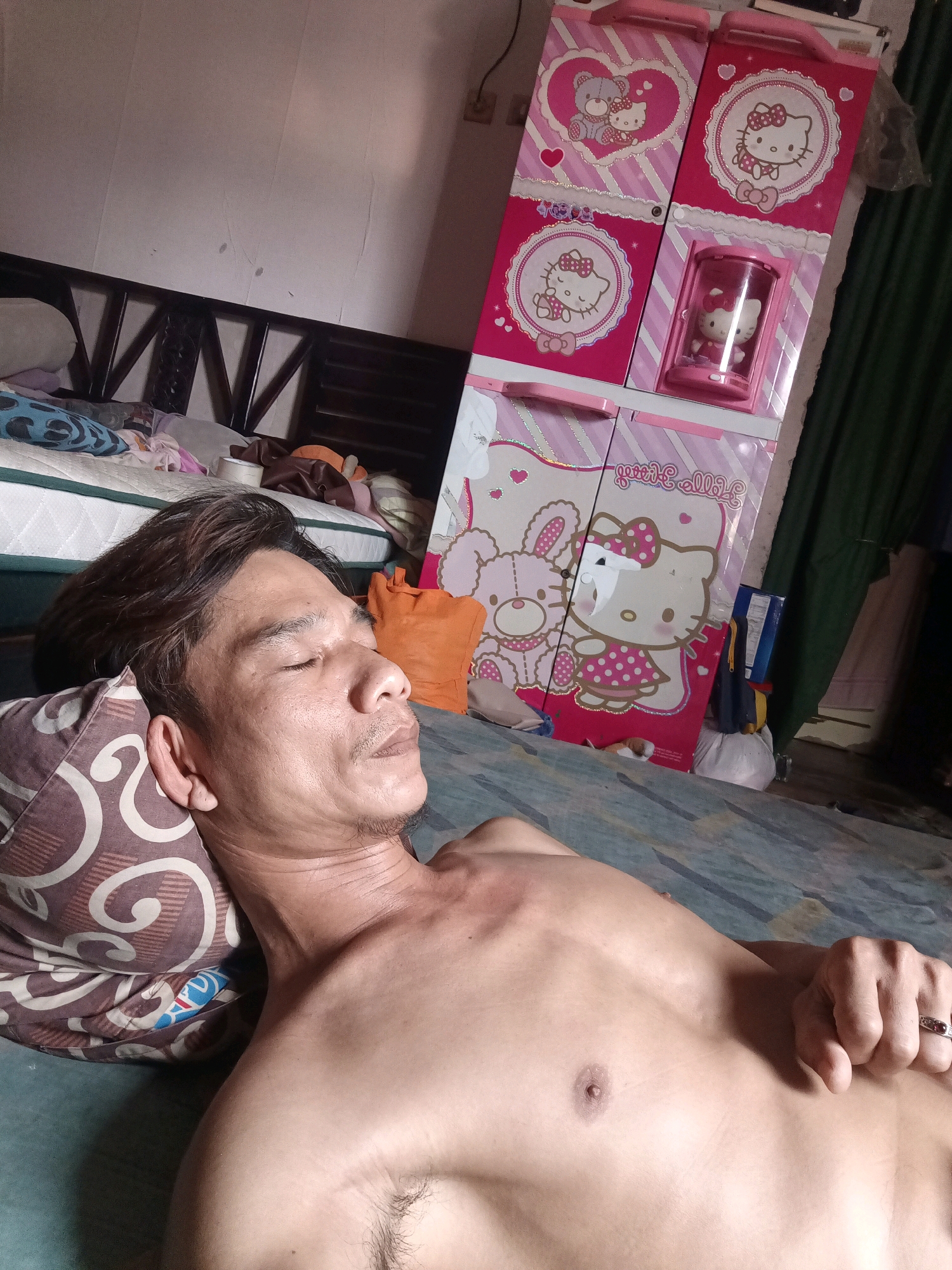A medium shot shows a man with his eyes closed, lying on his side on a patterned pillow in what appears to be a bedroom in South Jakarta, Indonesia. He is shirtless, and his left arm is resting across his chest, with his hand adorned with a ring. The background reveals a bed with a patterned mattress and various pillows, along with a tall pink cabinet decorated with Hello Kitty imagery. Other objects visible include what appears to be a mattress, a brown wooden headboard, and an orange pillow. The lighting suggests an indoor environment, possibly during the day or with artificial light.