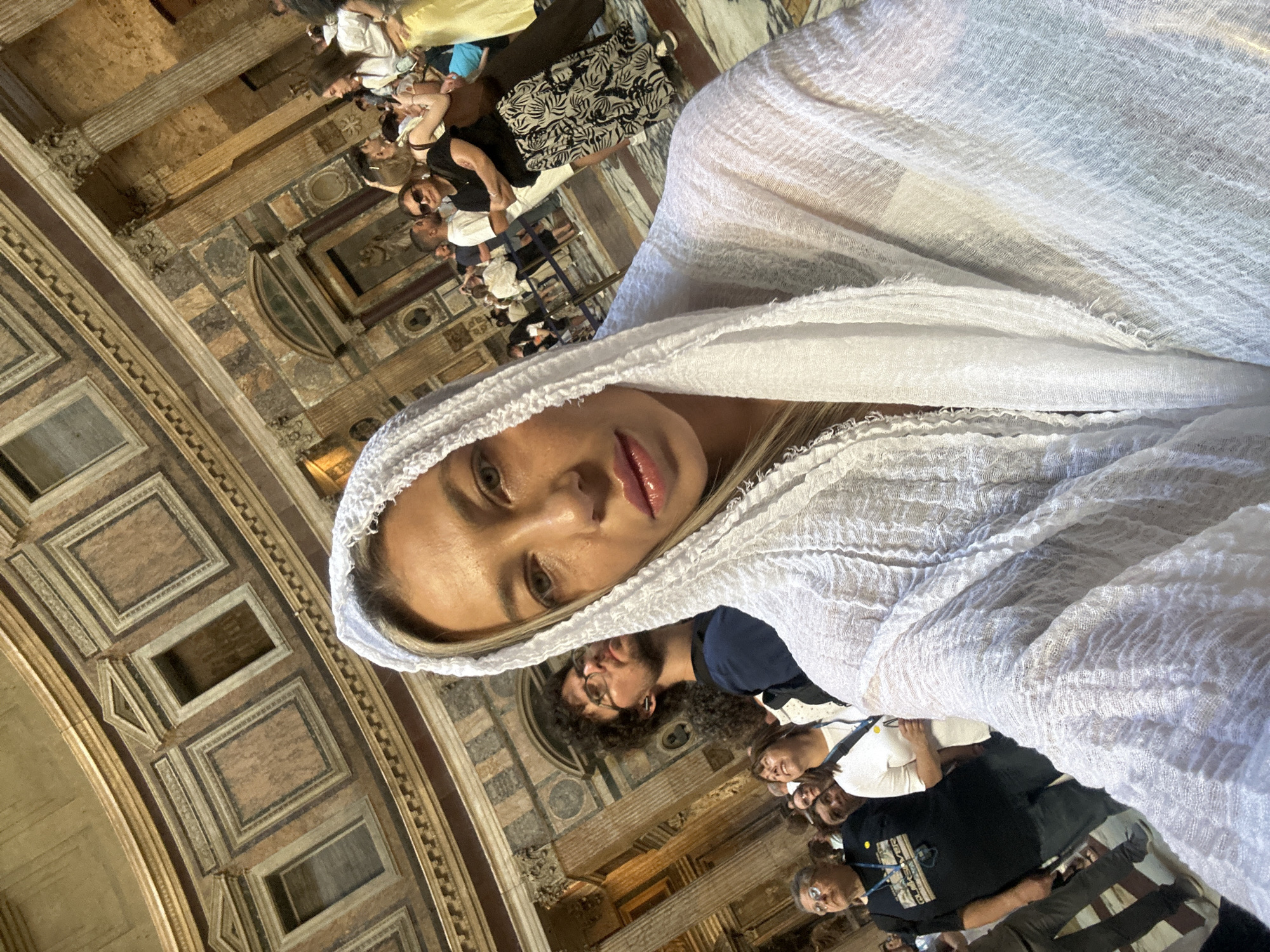 A woman wearing a white headscarf is posing for a selfie inside the Pantheon in Rome, Italy. Several other people are visible in the background. The Pantheon's interior architecture, including the coffered dome, is visible.