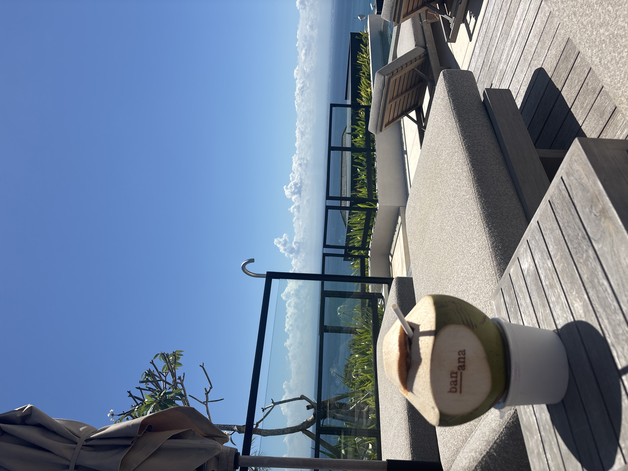 The image captures an outdoor scene in Kuta Selatan, Indonesia, featuring a coconut drink on a wooden surface, likely a table or lounge chair. The coconut, partially peeled and with a straw, sits in a white cup. The word "banana" is printed vertically on its surface. The foreground consists of wooden lounge furniture. In the background, a black metal and glass barrier separates the seating area from a view of the ocean and sky. The sky is predominantly blue with some white clouds on the horizon, near the ocean. Below the barrier, green foliage is visible. Partial visibility of a folded umbrella and plant life also appears. The overall lighting suggests a sunny day.