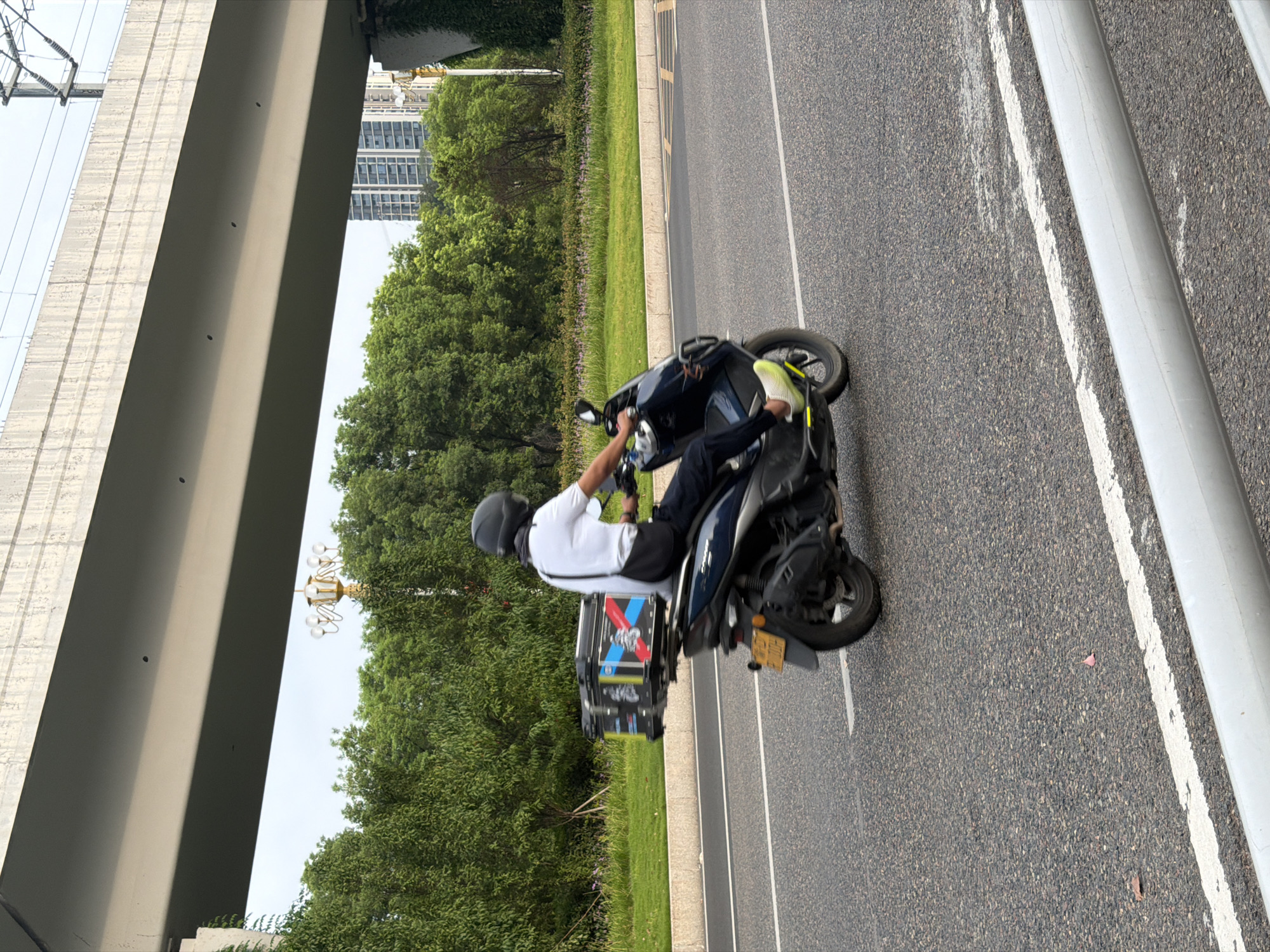 A person wearing a white shirt, dark pants, and a black helmet rides a dark blue scooter on a paved road under an elevated structure. The scooter has a black box mounted on the rear, adorned with a graphic featuring red and blue stripes and a motorcycle. The rider is partially obscured by the scooter and is wearing a dark bag slung across their body. The environment includes a grassy median with trees and bushes, a concrete elevated highway structure, and buildings in the distance. The road has white lane markings.