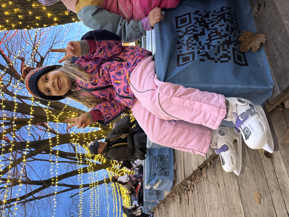 A young child, approximately six to eight years old, with blonde hair and wearing a multi-colored knit beanie, a pink leopard-print winter jacket, and pink snow pants, sits on a blue bench-like structure. The child is smiling and holding both hands up, forming "peace signs" with her fingers. She wears white ice skates with purple straps, labeled "Oxelo." To her immediate right, another child's arm and a portion of their pink and blue winter attire are visible. In the background, a man in a dark jacket and baseball cap sits on an identical blue bench, looking downwards. Several other indistinct figures are visible further back, also seated on similar benches. The benches are covered in a blue mesh material, featuring large black QR codes. The ground beneath consists of wooden planks with scattered brown autumn leaves. Overhead, numerous bare trees are densely draped with strings of warm yellow LED lights, creating an illuminated canopy against a deep blue twilight sky. The scene is located in an outdoor public area, likely an ice-skating rink changing or resting zone, in the city of Unknown, Unknown.