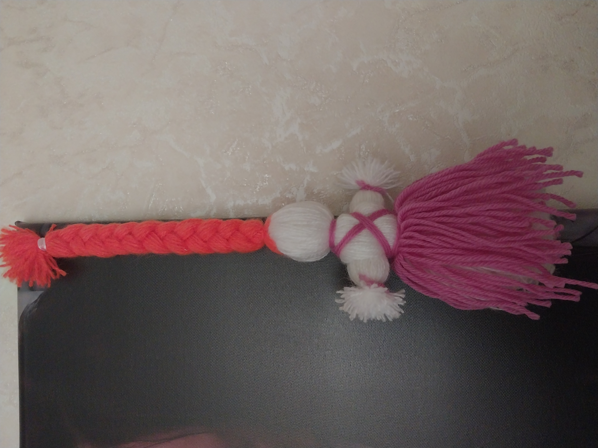 The image shows a close-up of a handmade yarn doll hanging from what appears to be a large framed picture or poster. The doll has bright orange yarn braided into a pigtail that hangs down. Below the pigtail is the doll's head, made of white yarn, and it has small white yarn pom-poms for arms. A pink yarn ribbon is wrapped around the doll's torso. The doll's skirt is made of densely packed strands of pink yarn. The background on the left is a dark, textured surface, possibly the framed picture itself, showing a hint of a person's face. The background on the right is a light-colored, textured wall, possibly wallpaper or painted plaster, with subtle marbling.
This image does not provide any information about its location in the city of Dnipro, Ukraine.
