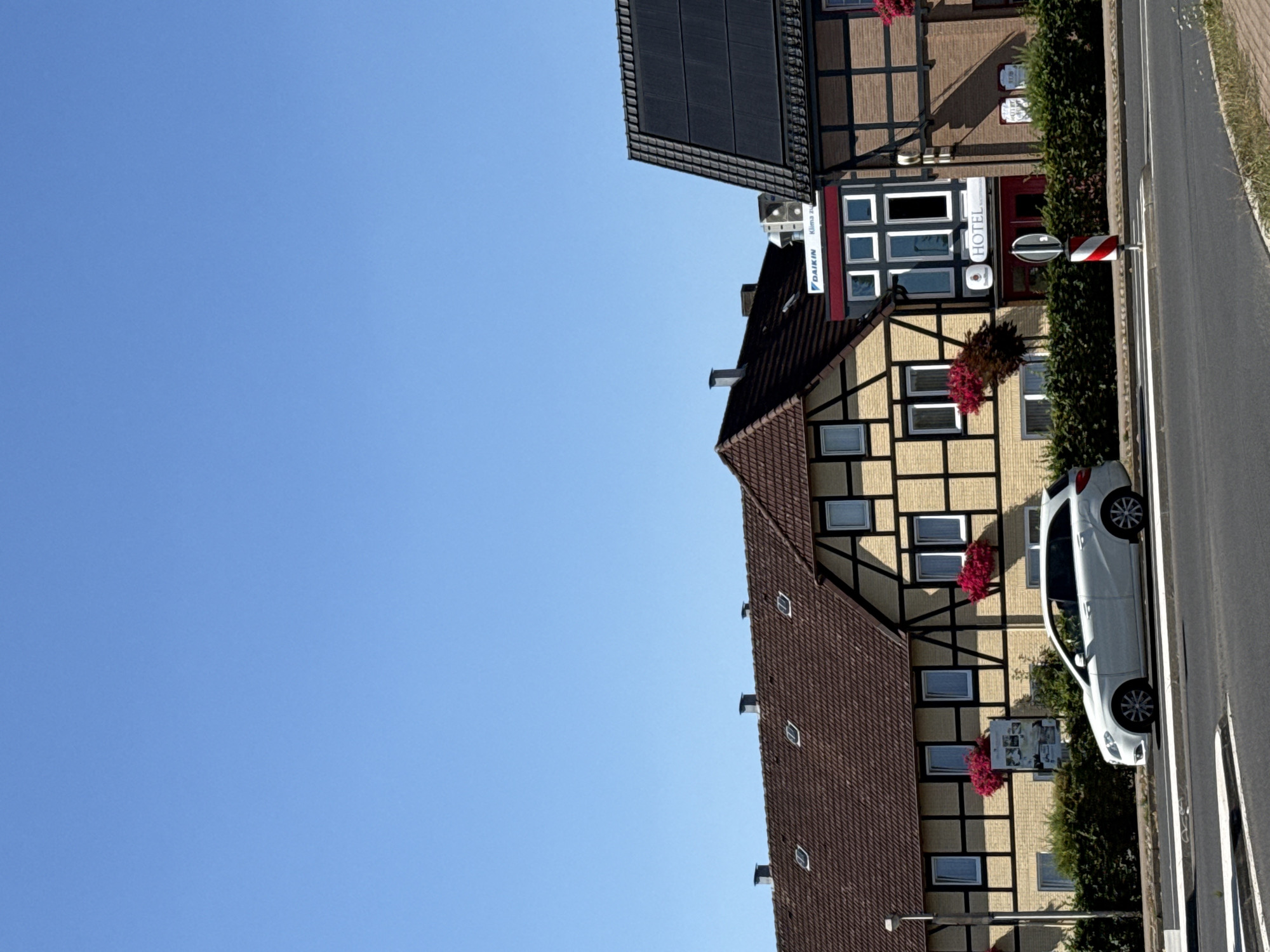 The image shows a street view in Herzberg, Germany. In the foreground, a white station wagon is parked on the side of the road. The car is facing right, and its headlights and taillights are visible. The road has a white line marking a crosswalk. Behind the car, a row of buildings lines the street. The buildings feature a traditional German architectural style with exposed timber frames and tiled roofs. Some of the buildings have signs indicating they are hotels. Red flowers are visible in window boxes on some of the buildings. The sky is clear and blue. The overall scene depicts a quiet street in a German town on a sunny day.