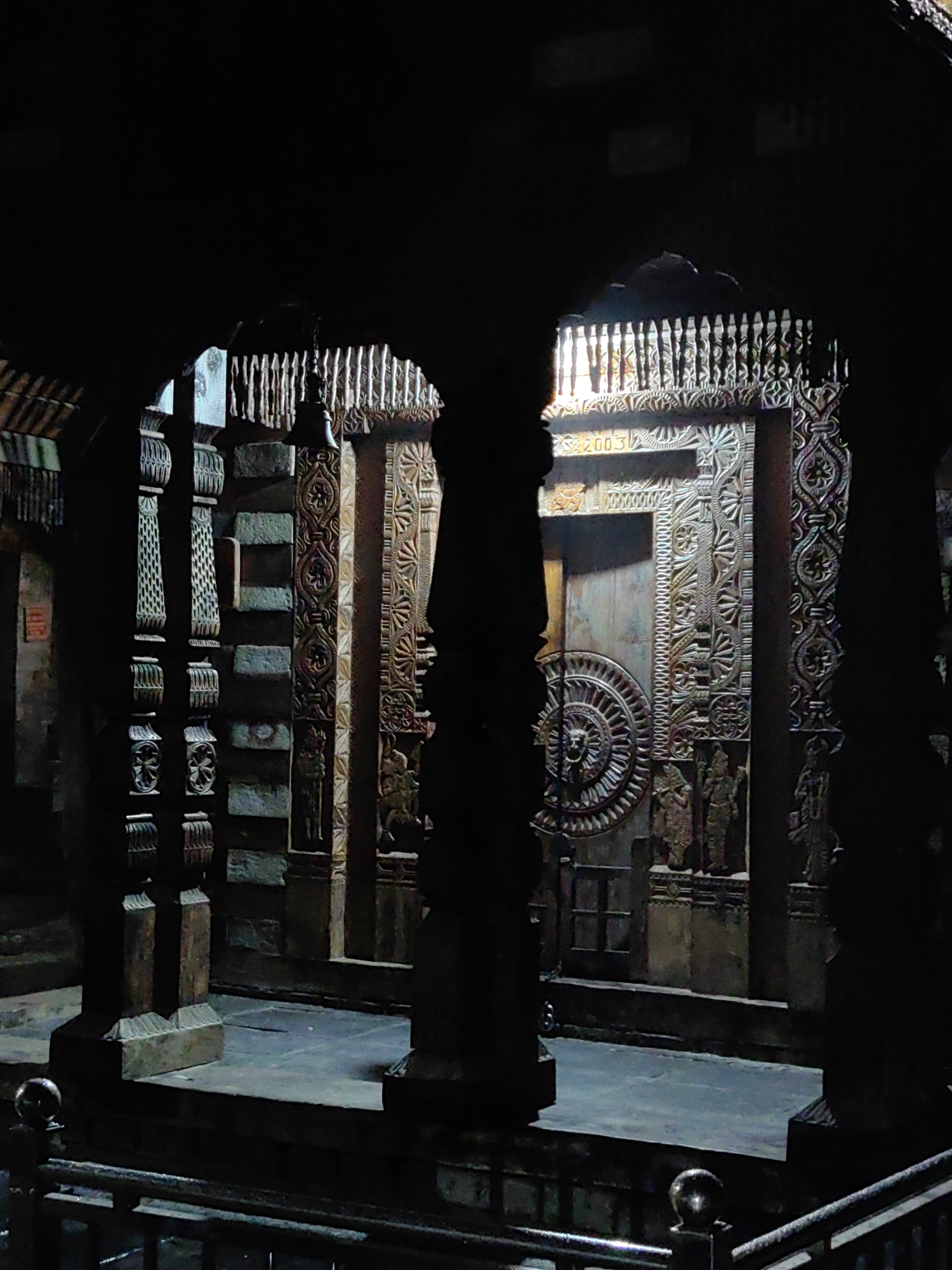 The image depicts the interior of a temple in Vashisht, India, characterized by its dark, dimly lit atmosphere and intricate architectural details. Prominently featured are carved wooden pillars and elaborately decorated walls, showcasing detailed traditional Indian motifs. In the background, a large, circular carving with a face-like center is visible, flanked by smaller carved figures of deities or historical personages. The overall impression is one of ancient religious artistry and a somber, reverent ambiance. The lighting is dramatic, with light sources illuminating specific areas, creating strong contrasts between light and shadow, which enhances the textures and carvings. The scene appears to be uninhabited, with no people visible in the frame.