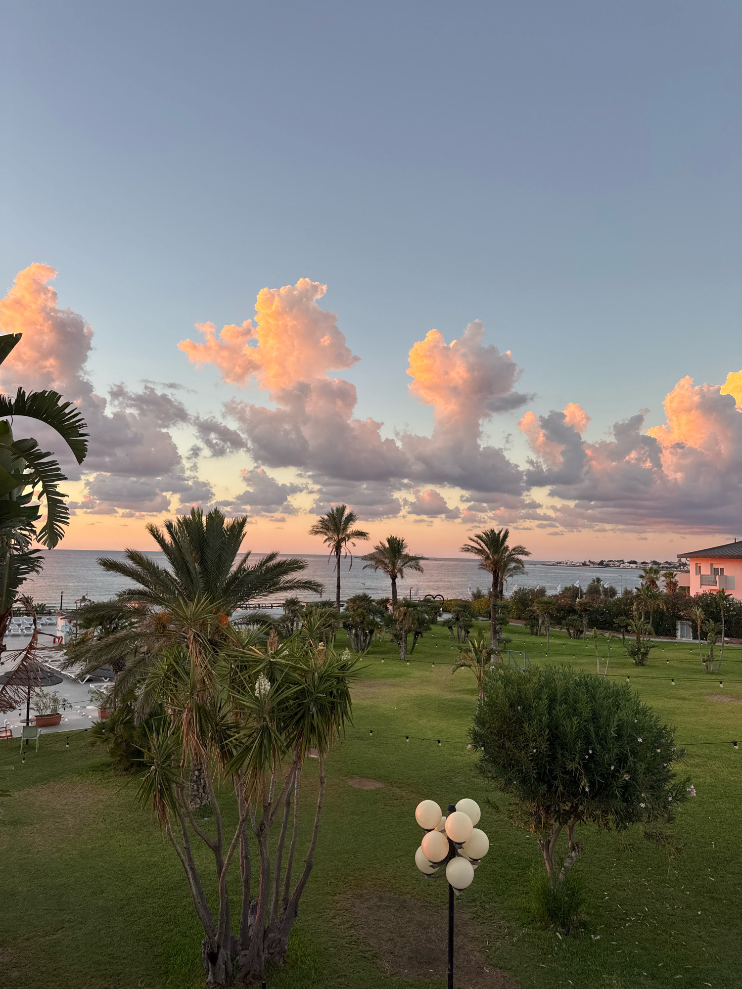 This image depicts a sunset view over the Mediterranean Sea in Isola delle Femmine, Italy. The sky is a gradient of blue and pink, with fluffy clouds catching the warm hues of the setting sun. Palm trees and other lush vegetation dominate the foreground, with a well-manicured lawn stretching towards the sea. In the distance, a pink building with a dark roof is visible, suggesting a hotel or resort. The sea is calm, with a few boats docked along the shore. The overall atmosphere is serene and picturesque, characteristic of a coastal Italian town. There are no people or animals visible in the frame.