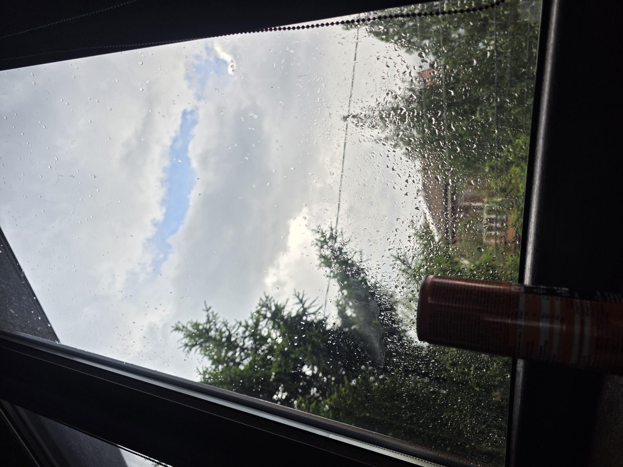 The image shows a view through a rain-streaked window, likely a sunroof or car window. Outside, the sky is overcast with patches of blue. Green trees are visible, some blurred by the rain on the glass, and a building with a reddish roof can be seen in the background. An aerosol can with a red and white label rests against the window frame on the right side. The location is unknown, there is no explicit evidence that this image was taken in the city of Yagodovo, Bulgaria.