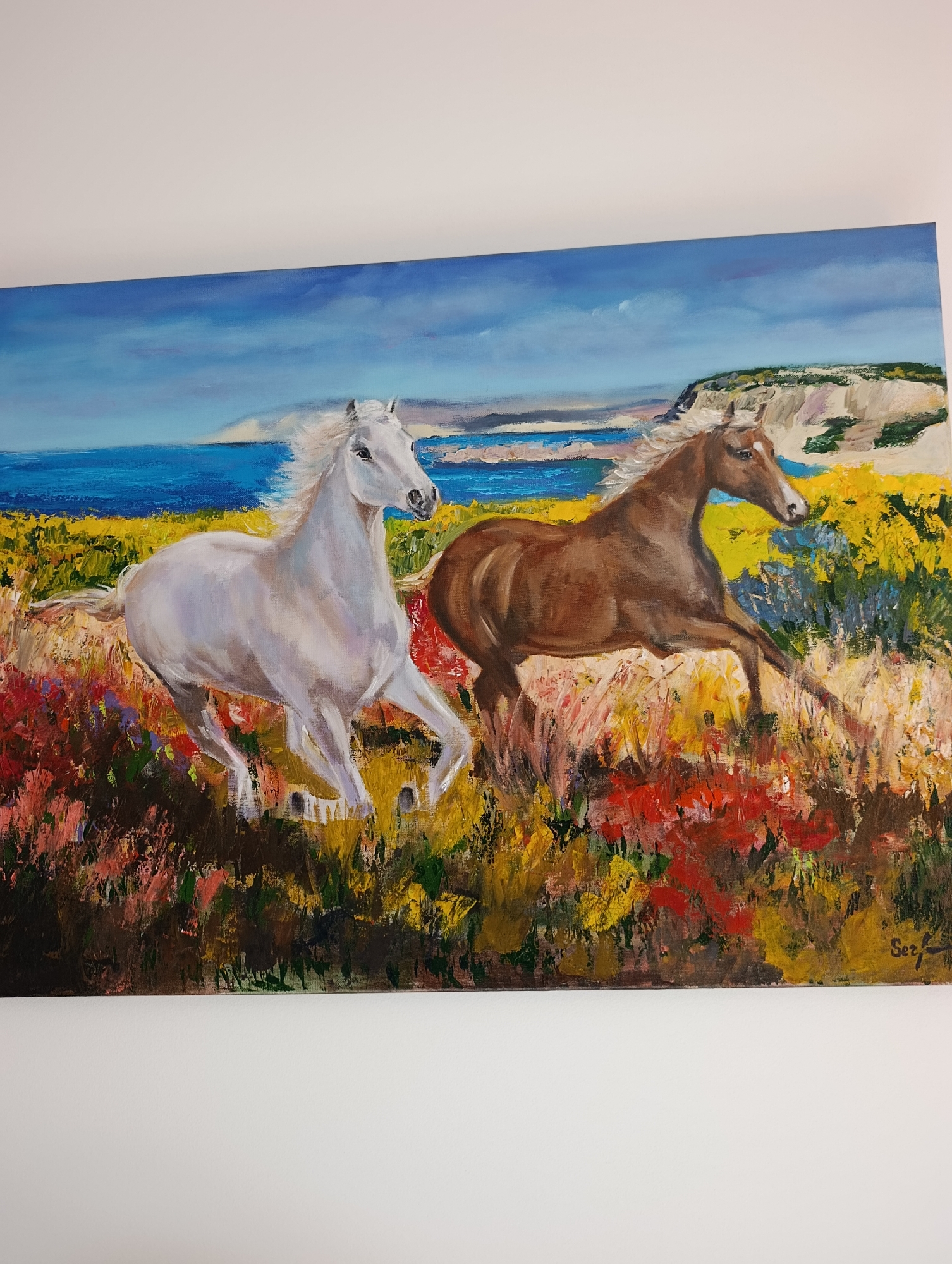 The image displays a painting of two horses running through a field of colorful flowers with a body of water and a distant landscape in the background. The horse on the left is white and the horse on the right is brown. The sky is a vibrant blue with some clouds. The painting is signed in the lower right corner with "Ser". The location in Setúbal, Portugal is not discernable from the image itself.