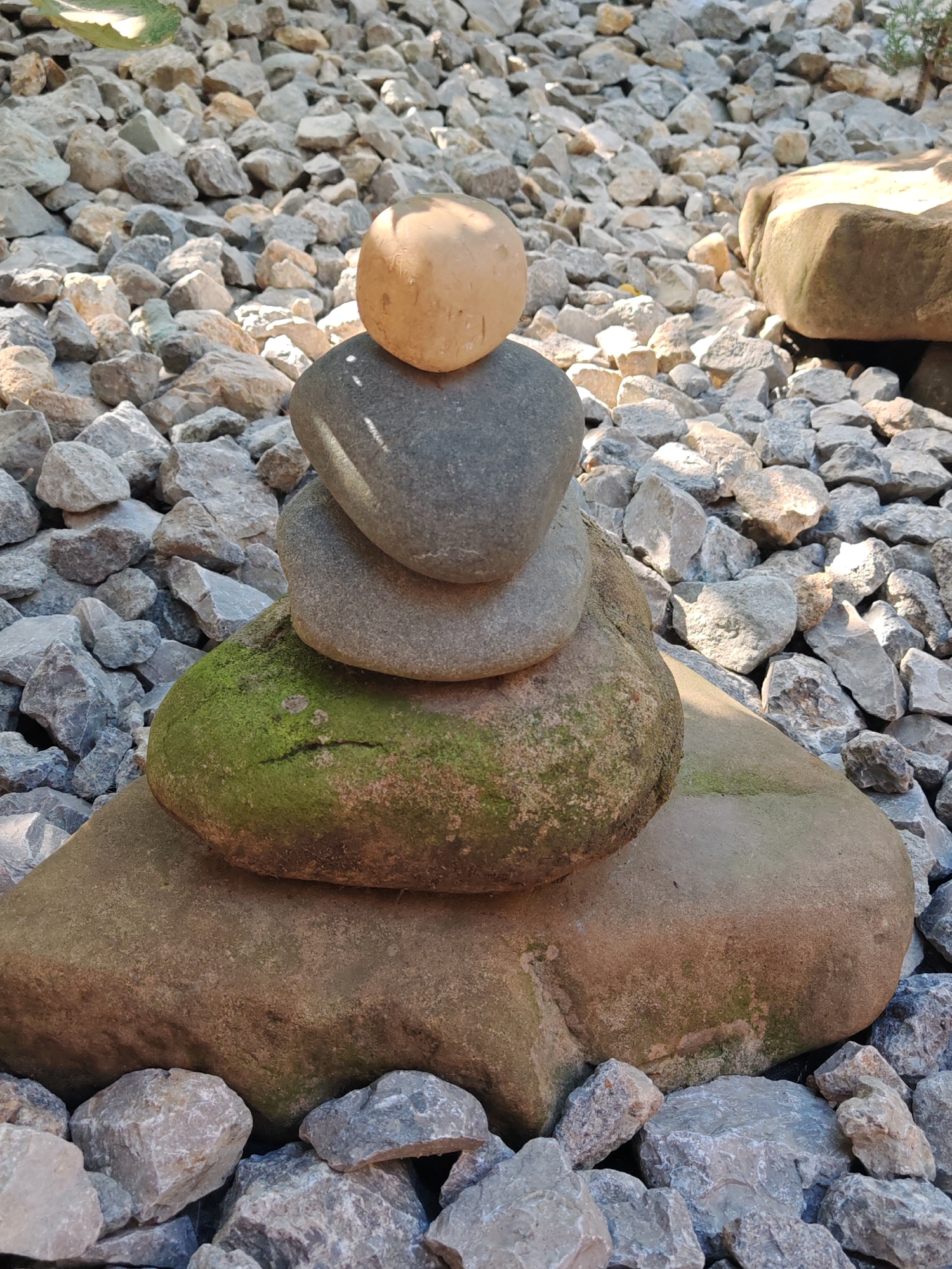 A cairn of five stones is stacked on a bed of gravel in a sunny outdoor setting. The bottom stone is large and flat, with a textured surface. The second stone from the bottom is partially covered in green moss. The third and fourth stones are smaller and smooth, with greyish tones. The top stone is a rounded, light brown pebble. The surrounding gravel consists of various sizes of grey, tan, and white stones, with some larger, light brown rocks visible in the background. The lighting suggests it is daytime.