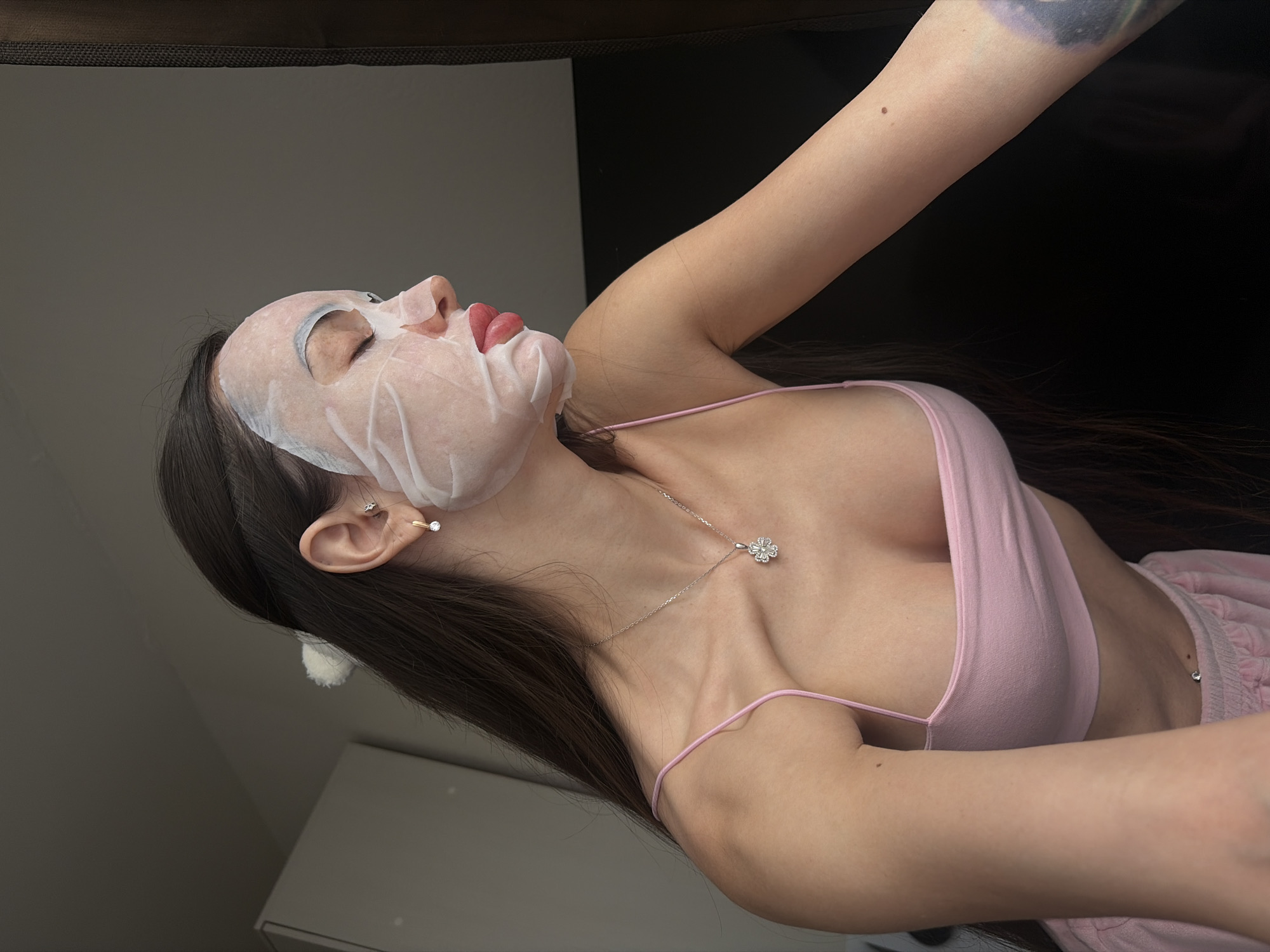 A young woman lies on her back, wearing a pink tank top and pajama bottoms. She has a white sheet mask on her face. She also wears earrings and a necklace. The background is a mix of light grey and black.