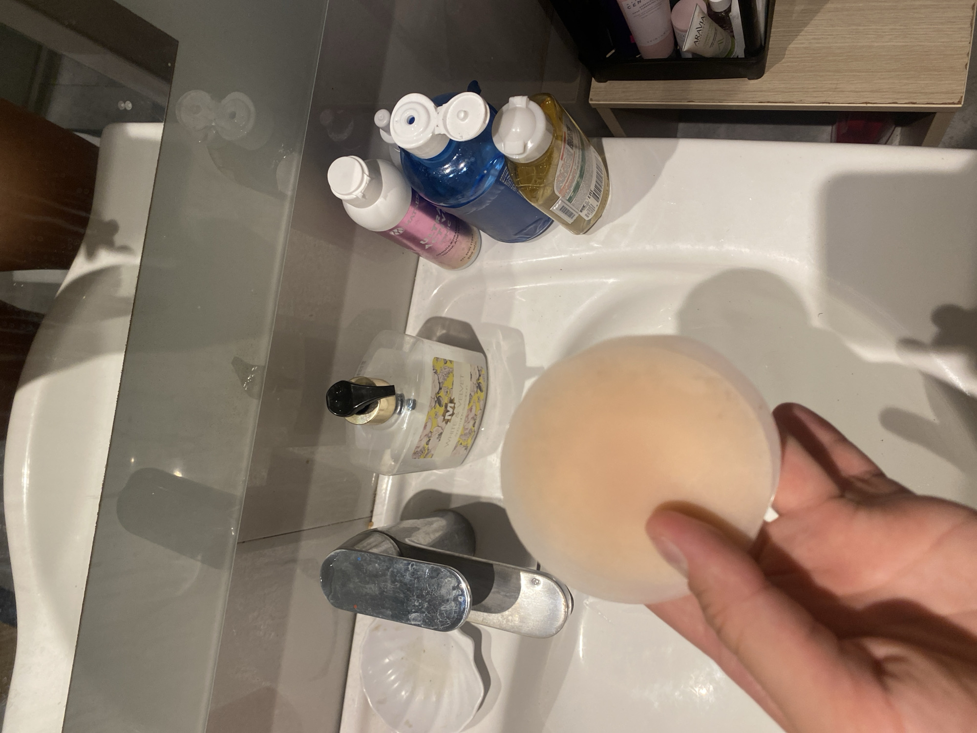 The image shows a bathroom sink area with various items. In the foreground, a hand is holding a flesh-toned nipple cover. To the left of the hand is a chrome faucet. Behind the faucet is a white, shell-shaped dish. Further back, on the sink, is a bottle of "WHITE VELVET" hand soap. To the right of the soap are several other bottles, including a pink bottle, a blue bottle, and a yellow bottle. In the background, a mirror reflects the ceiling and a portion of a person's legs and torso. A shelf unit with cosmetic products is visible on the far right. The scene appears to be indoors, in a domestic bathroom setting. There is no discernible information in the image that places it in Aksu, Turkey.