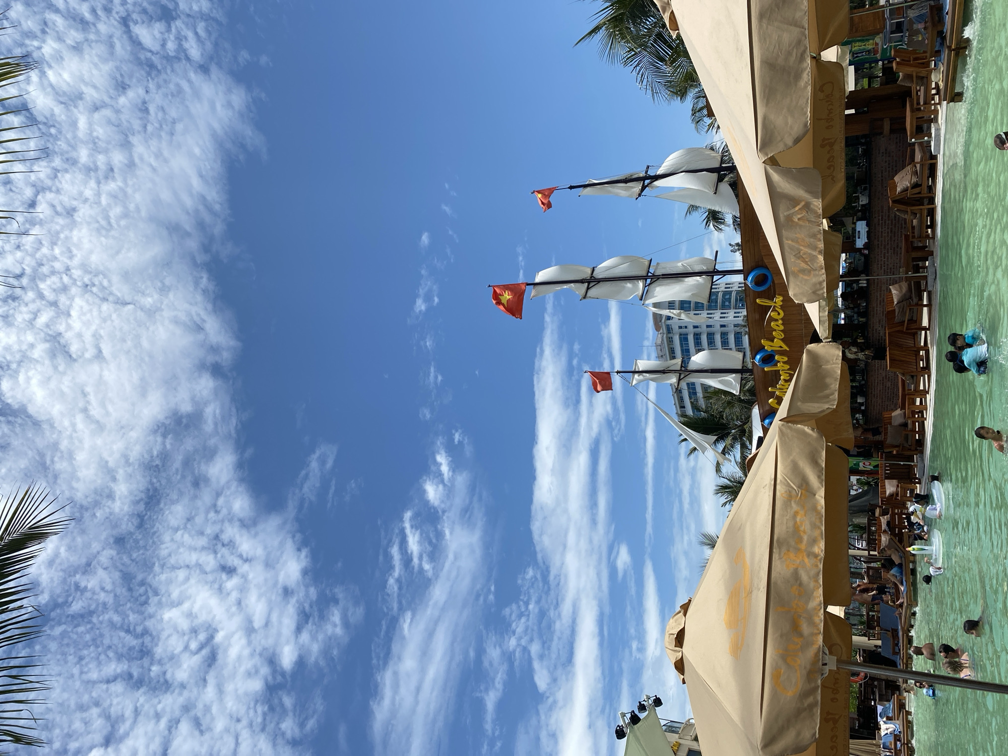 The image captures a sunny day at the beachside of Da Nang, Vietnam, likely at a resort or entertainment complex. In the foreground, large beige umbrellas with the text "Golden Beach" are prominent, providing shade over a shallow, turquoise swimming pool. Several people are visible in the pool, including adults and children, some swimming and others relaxing. The pool area is furnished with lounge chairs and tables, suggesting a leisure and entertainment venue.
In the mid-ground, a bar area with wooden furniture and brick accents is partially visible behind the umbrellas. Behind the bar, a large, decorative pirate ship with white sails and red flags stands out against the blue sky. The ship's masts are adorned with Vietnamese flags. In the background, a modern, multi-story building is visible, suggesting the proximity of a hotel or other urban structures.
The sky is a clear blue with scattered white clouds, and palm fronds are visible at the top of the frame, indicating a tropical or subtropical location. The overall scene conveys a vibrant atmosphere of a popular tourist destination in Da Nang, likely during the daytime.