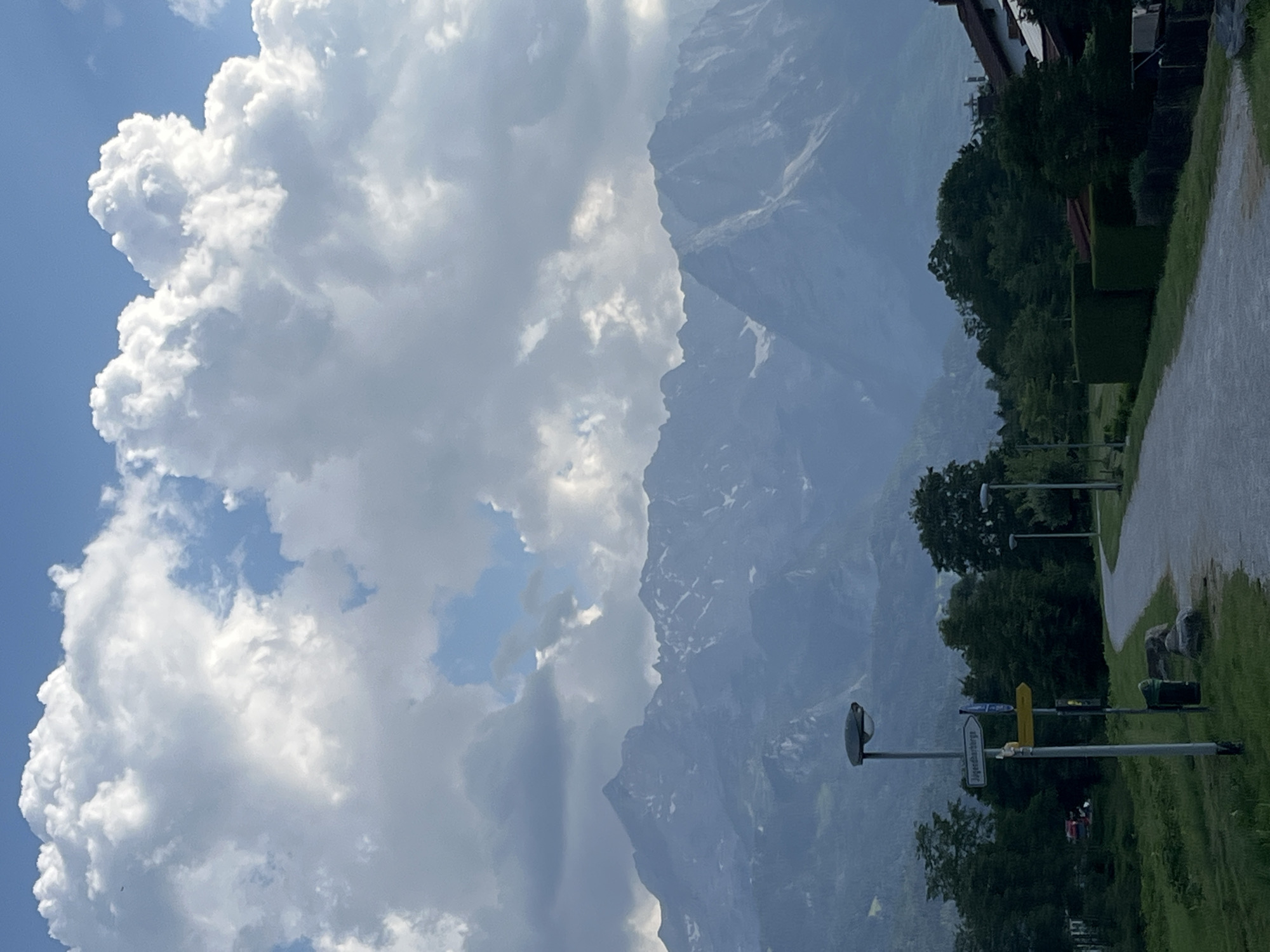 The image captures a scenic view of the Bavarian Alps, likely from the outskirts of Garmisch-Partenkirchen, Germany. In the foreground, a paved path curves through a grassy area with trees lining its sides. A lamppost with a signpost showing directions stands to the left of the path. Beyond the trees, a range of majestic, snow-capped mountains dominates the background, partially obscured by dramatic, billowing clouds. The sky is a mix of blue and grey, with the sunlight filtering through the clouds. The scene appears to be an outdoor recreational area or a scenic route, possibly a hiking or cycling path, leading towards the imposing mountain landscape. There are no people or animals visible in the image. The overall atmosphere is one of natural beauty and tranquility.